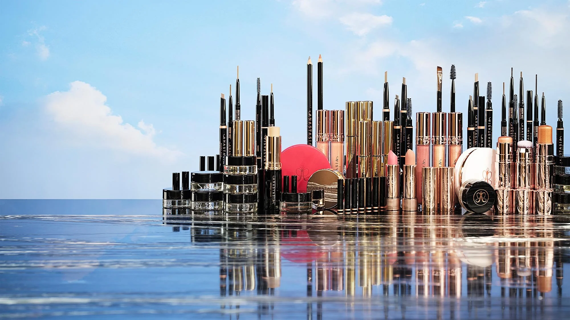 A collection of makeup products, including lipsticks, eyeliners, and brushes, arranged on a reflective surface outdoors with a blue sky and clouds in the background.