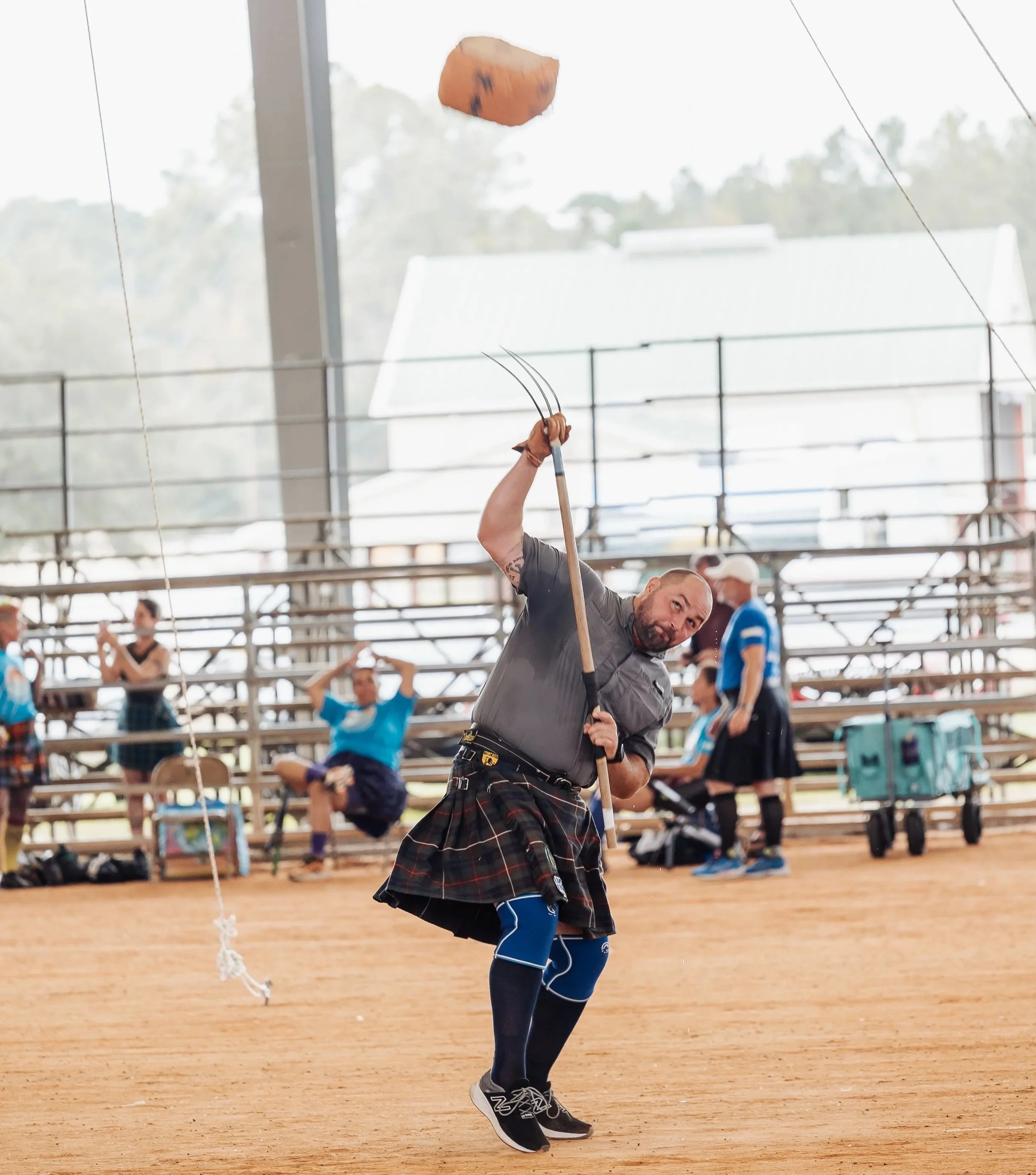 Man in traditional Scottish attire swinging a stick at a haggis during an event, with spectators sitting in bleachers in the background.
