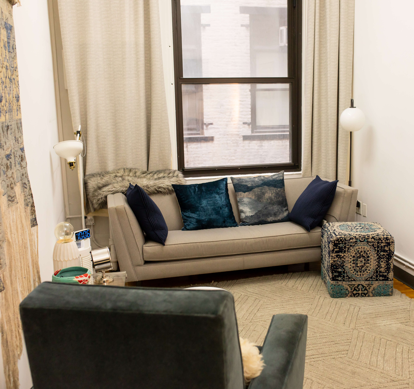 A therapy room with a beige sofa decorated with dark blue pillows, a small patterned ottoman, and a wall-mounted lamp. There are curtains on a window showing an urban exterior.