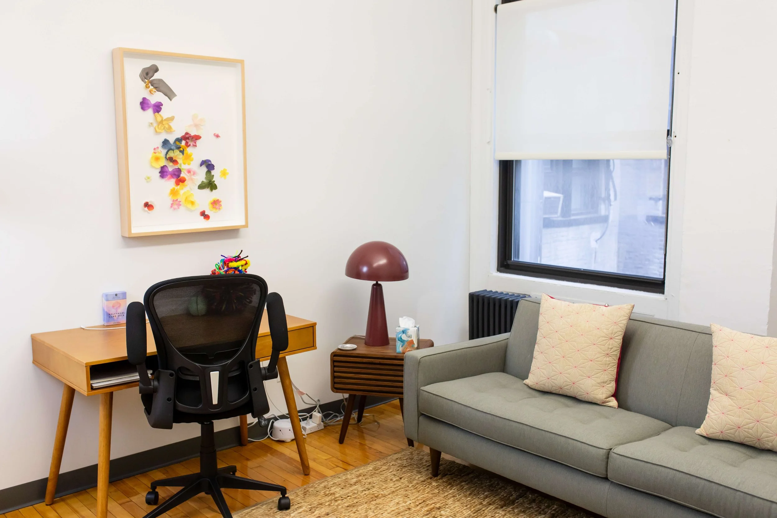 A cozy office corner with a wooden desk and black ergonomic chair, a small side table with a vintage-style lamp, a gray sofa with patterned pillows, a window with a white blind, and a wall art piece with colorful felt decorations.