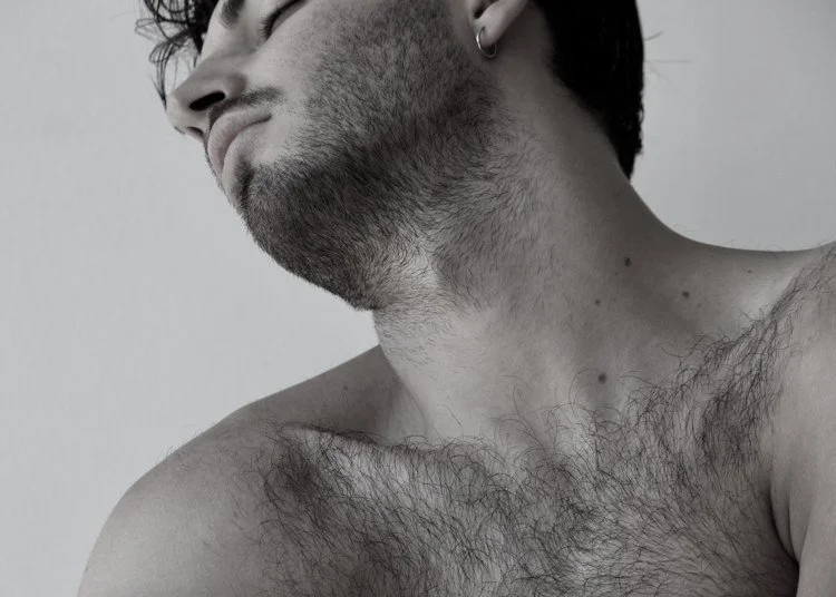 Close-up of a shirtless man with facial hair, wearing an earring, looking to the side against a plain background.