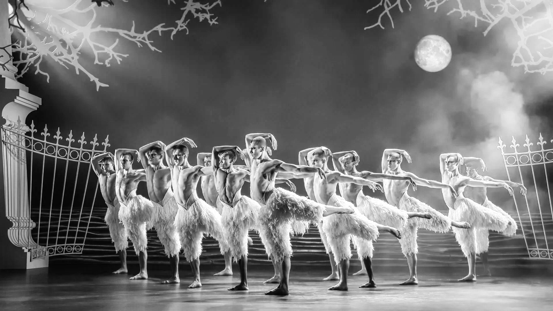 Group of ballet dancers performing on stage outdoors under moonlight, dressed in feathered costumes, with a decorative gate and large moon in the background, in black and white.