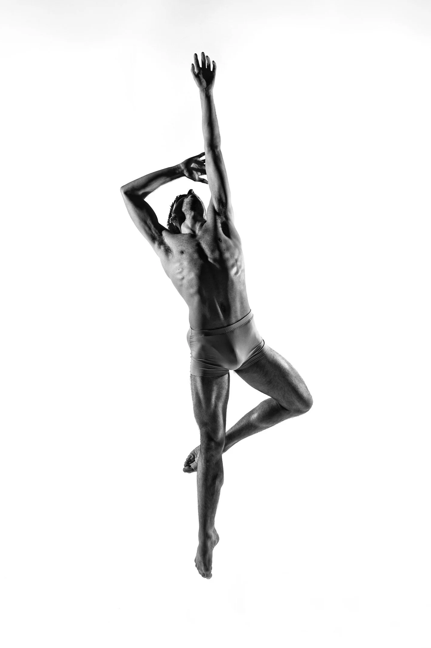 A black and white photo of a male ballet dancer in an artistic pose, standing on one leg with the other knee bent, one arm reaching upward and the other bent near his face.