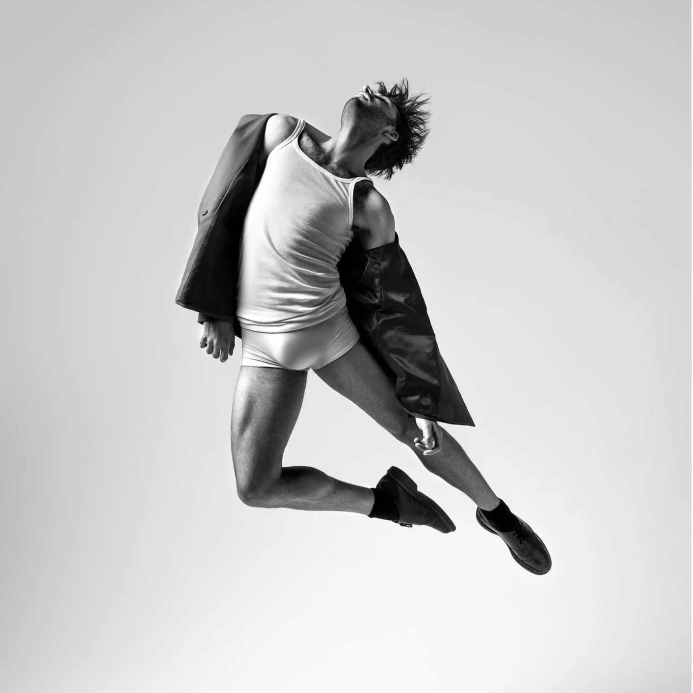 Black and white photo of a man in mid-air with long hair, wearing a tank top, shorts, a jacket draped over his shoulders, and dress shoes, appearing joyful or expressive.