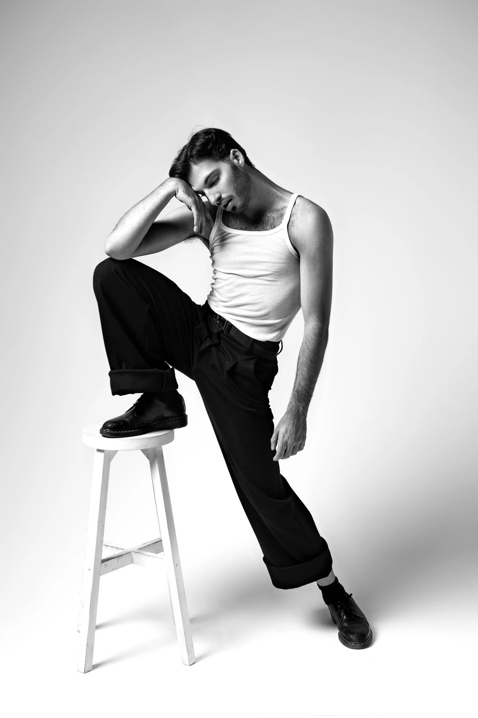 A black and white photo of a man posing with one foot on a stool, wearing a white tank top, dark pants, and black shoes, with a contemplative expression and his head resting on his hand.