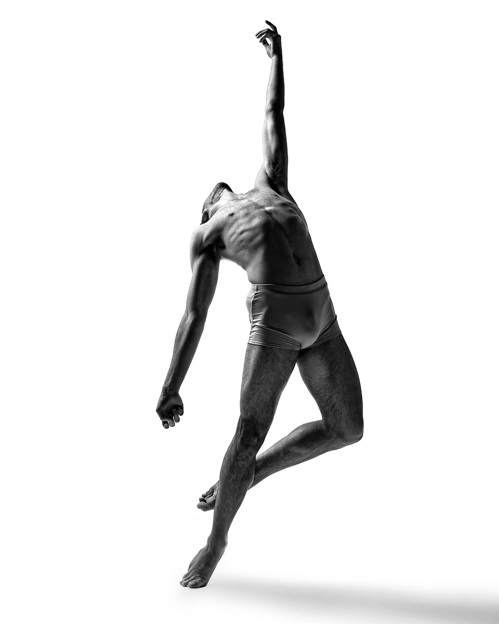 A black-and-white image of a muscular man in underwear performing a dance or pose with one arm extended upward and the other arm relaxed downward.