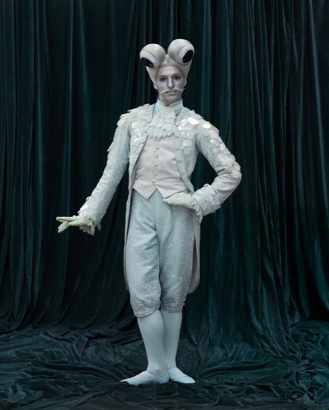 Person dressed in elaborate costume resembling a fantasy character, with large eye-shaped headpiece, light-colored clothing, and standing on a dark fabric backdrop.