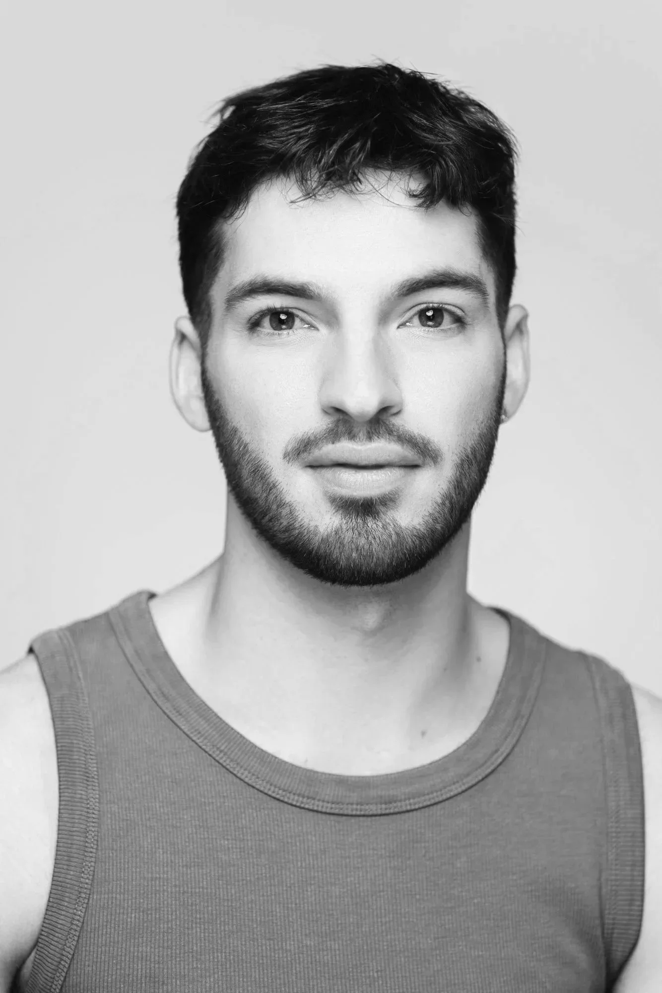 Black and white portrait of a young man with short dark hair, a beard, and mustache, wearing a sleeveless shirt, facing forward with neutral expression.