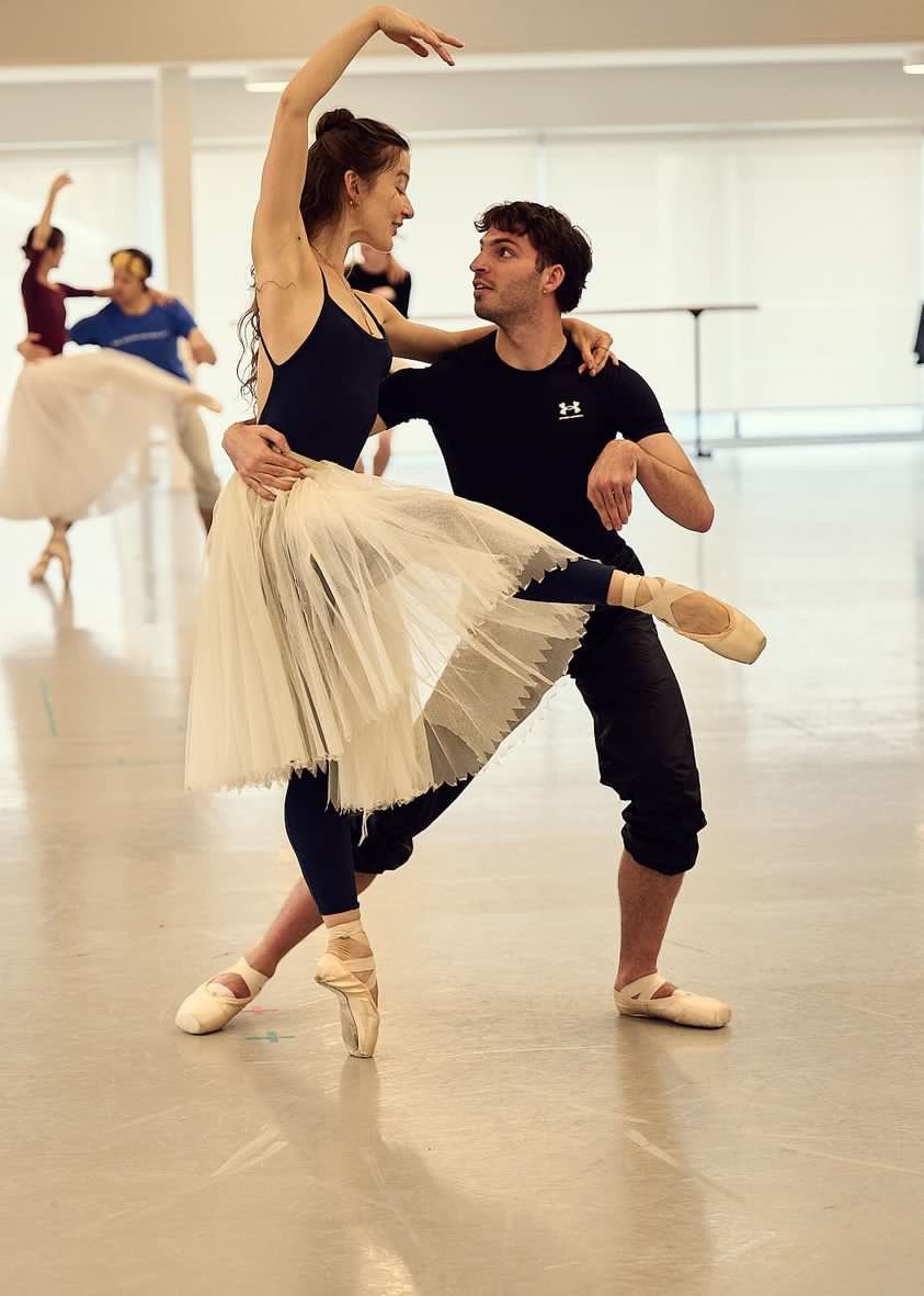 A ballet dancer and male ballet instructor practicing in a dance studio, with other dancers in the background.