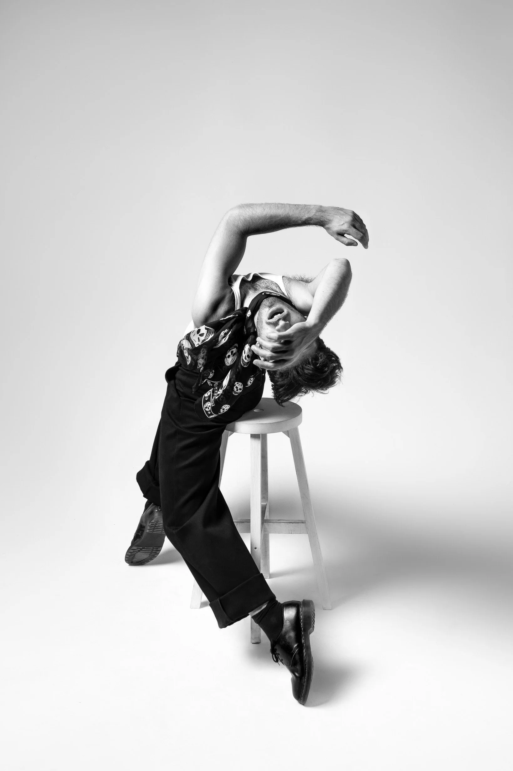 Black and white photo of a person leaning backwards with their head on a stool, one hand covering their face, wearing a bandana and black shoes, in a studio setting.