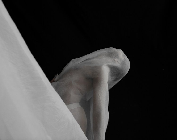 A close-up of a human figure covered in white fabric, sitting with a dark background.