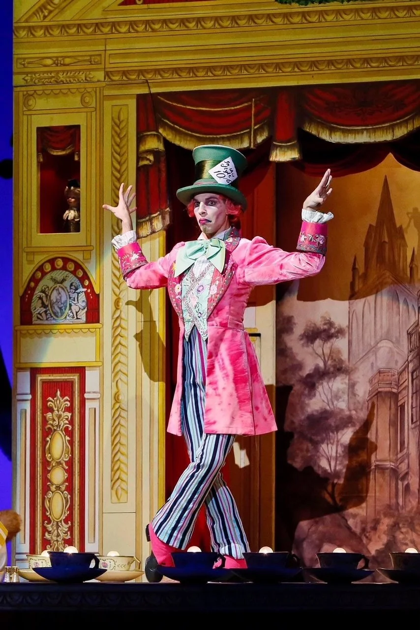 Costumed performer dressed as the Mad Hatter from Alice in Wonderland standing on stage with arms raised, in front of a theatrical set resembling a palace or castle.