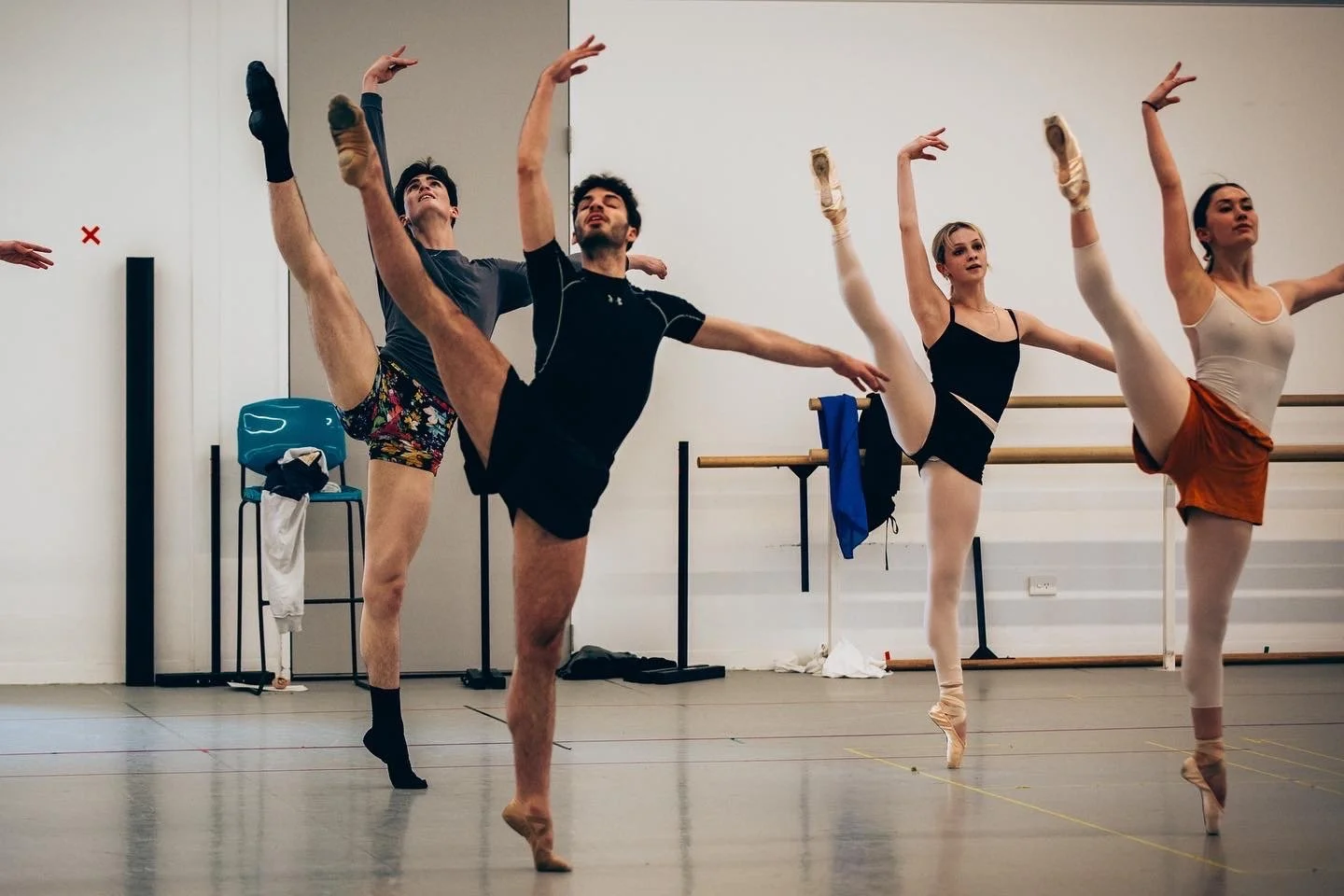 Ballet dancers practicing in a dance studio, with one male dancer lifting a leg high while others hold poses at the barre.