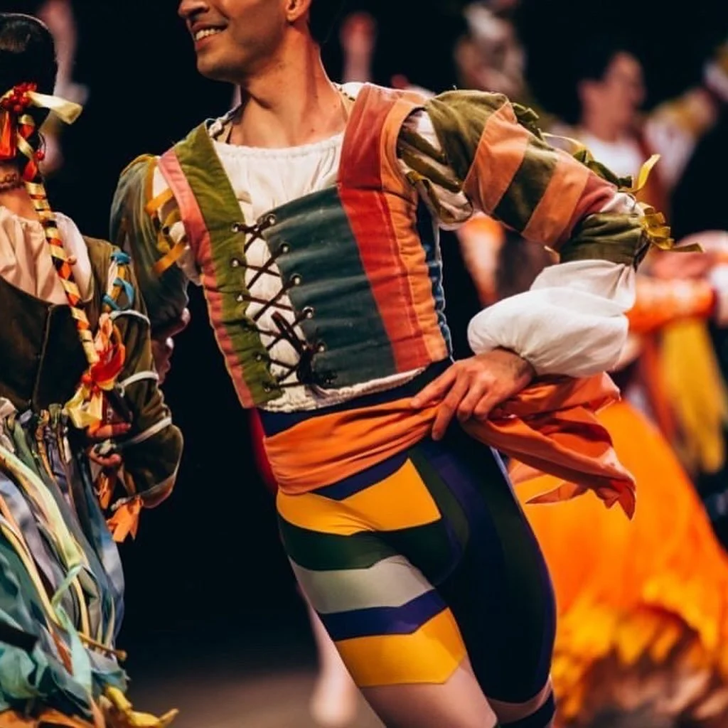 A man dressed in a colorful, traditional costume, smiling at a cultural event or festival, with other similarly dressed people in the background.