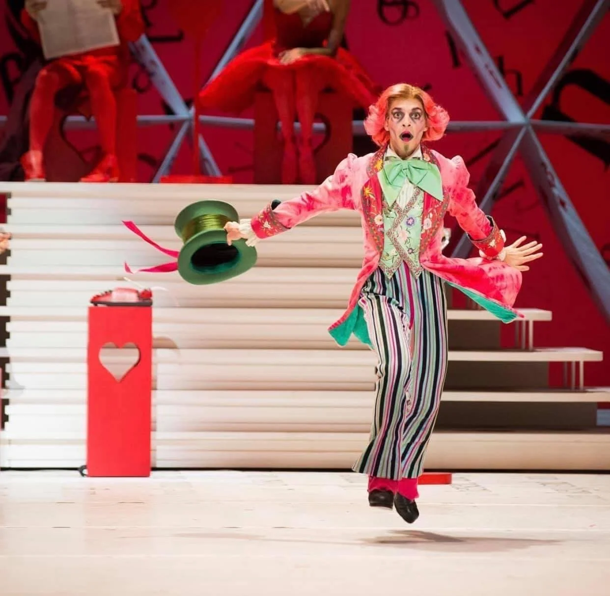 A performer dressed in a colorful, eccentric costume, including striped pants and a pink jacket with floral patterns, jumps in the air with a surprised expression and outstretched arms. The background features a red stage set with white stairs, a red