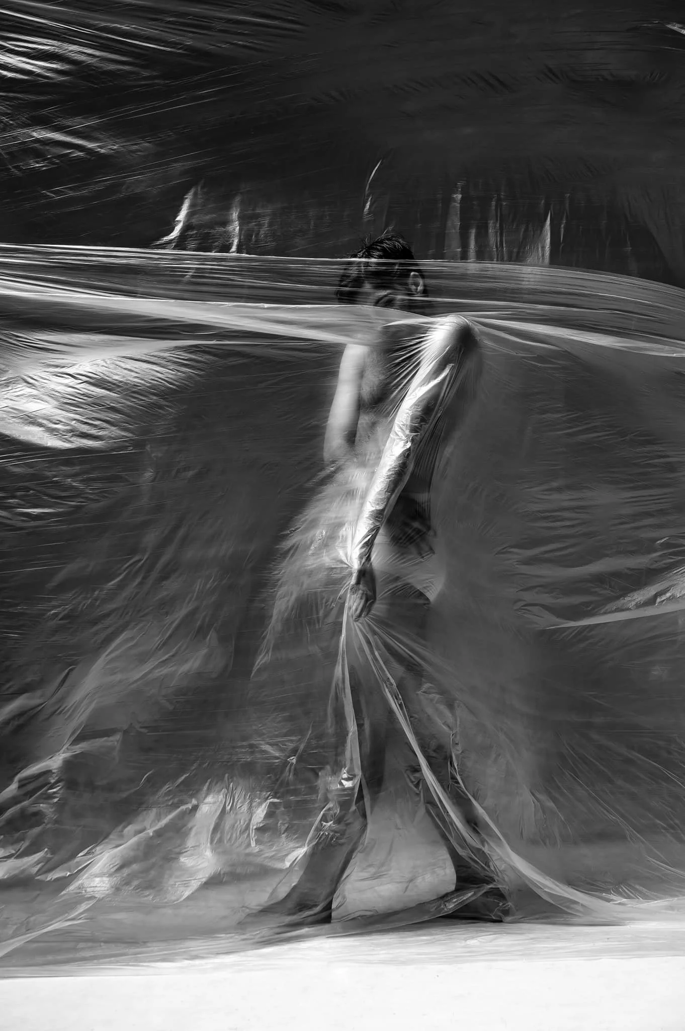 A person is walking behind a semi-transparent plastic sheet in a black-and-white photo, obscuring most of their body and face.
