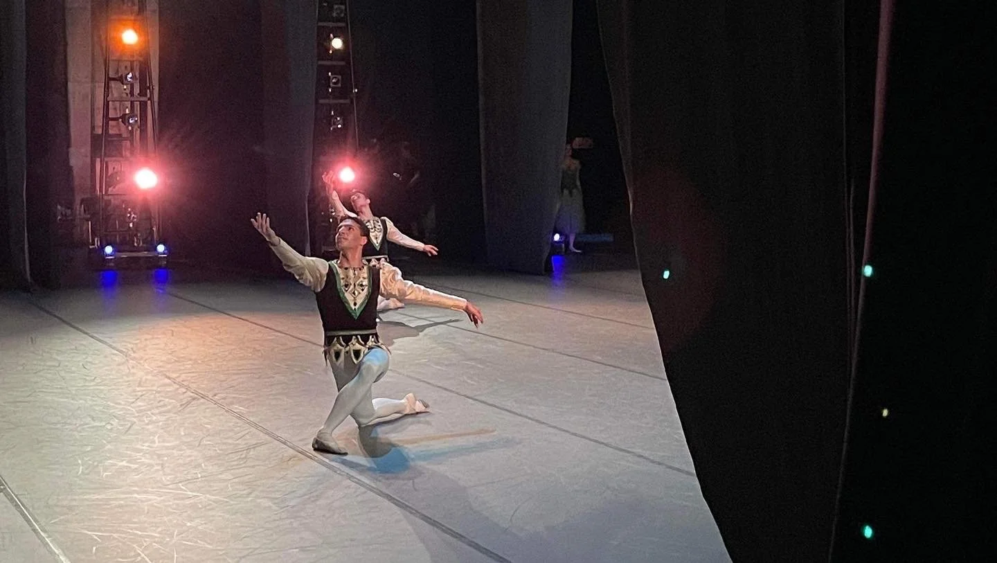 Two ballet dancers wearing traditional costumes perform on stage, one kneeling with arms extended and the other standing with arms raised. Stage lighting with red, orange, and purple lights in the background.