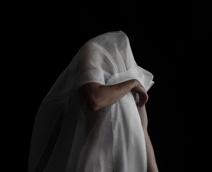 Person draped in a sheer white fabric against a black background, partially covering their face and body.