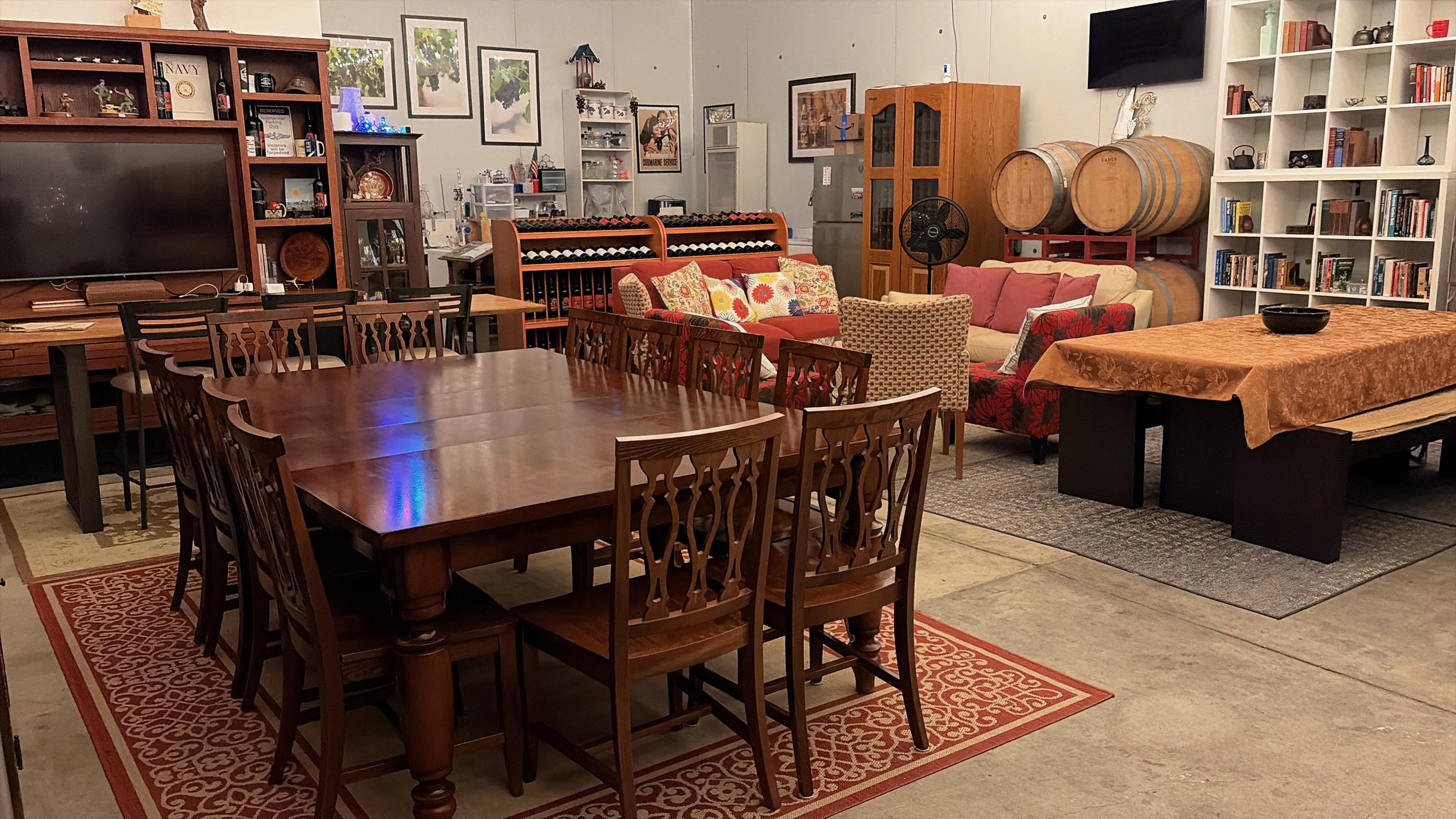 A cozy room with a large wooden dining table surrounded by wooden chairs and a multicolored area rug underneath. In the background, there is a seating area with different sofas and armchairs, some with floral cushions. Behind the seating area, shelves filled with books, decorative items, and two large wine barrels are visible. The room has artwork on the walls and various decorative items on the shelves.