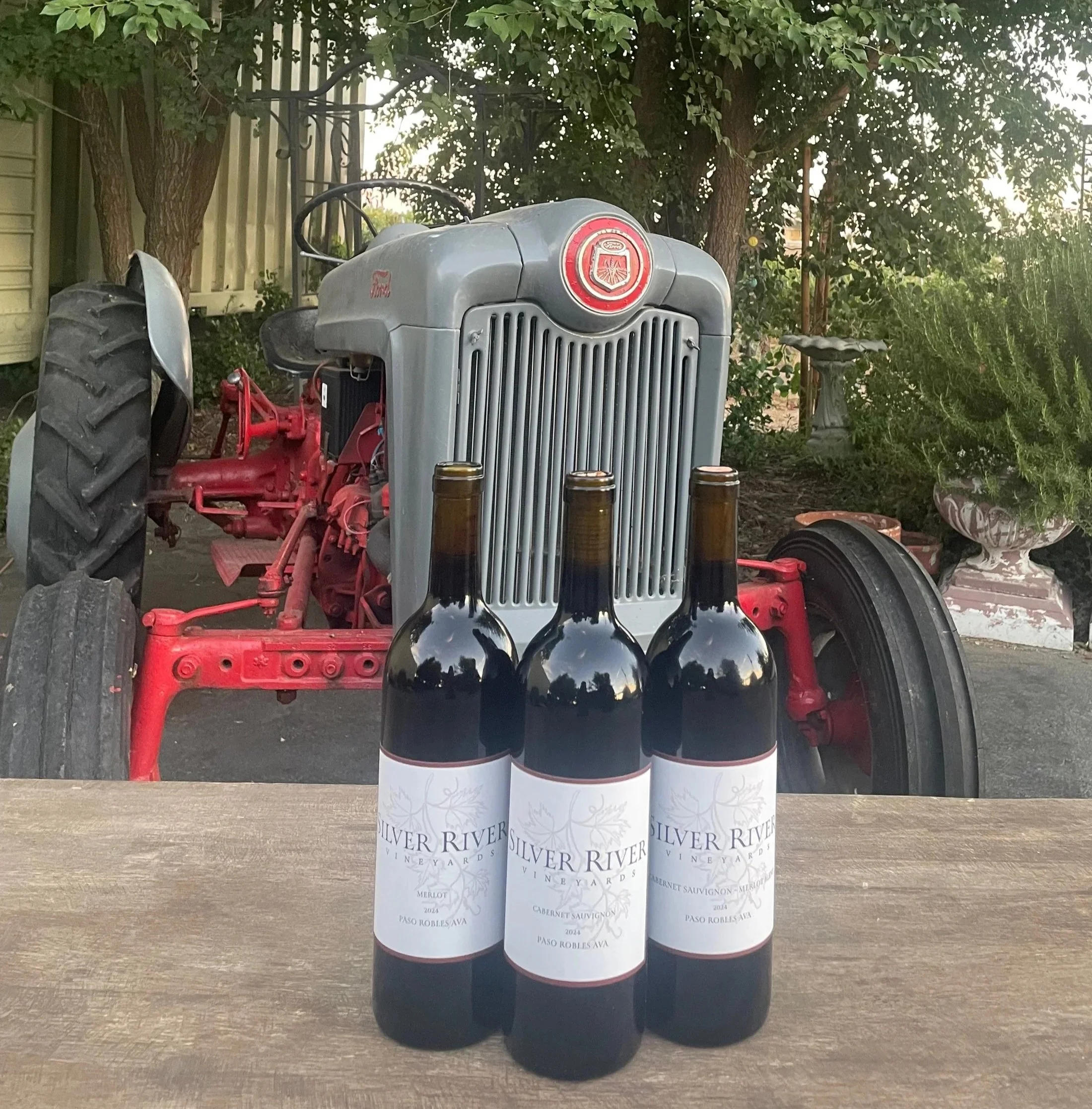 Four bottles of Silver River Vineyards wine are placed on a wooden surface outdoors, with three black top wine bottles and one white at the bottom. The wine bottle labels display the vineyard's name and wine types, such as Merlot, Cabernet Sauvignon, and D'Auge, along with vintage years.