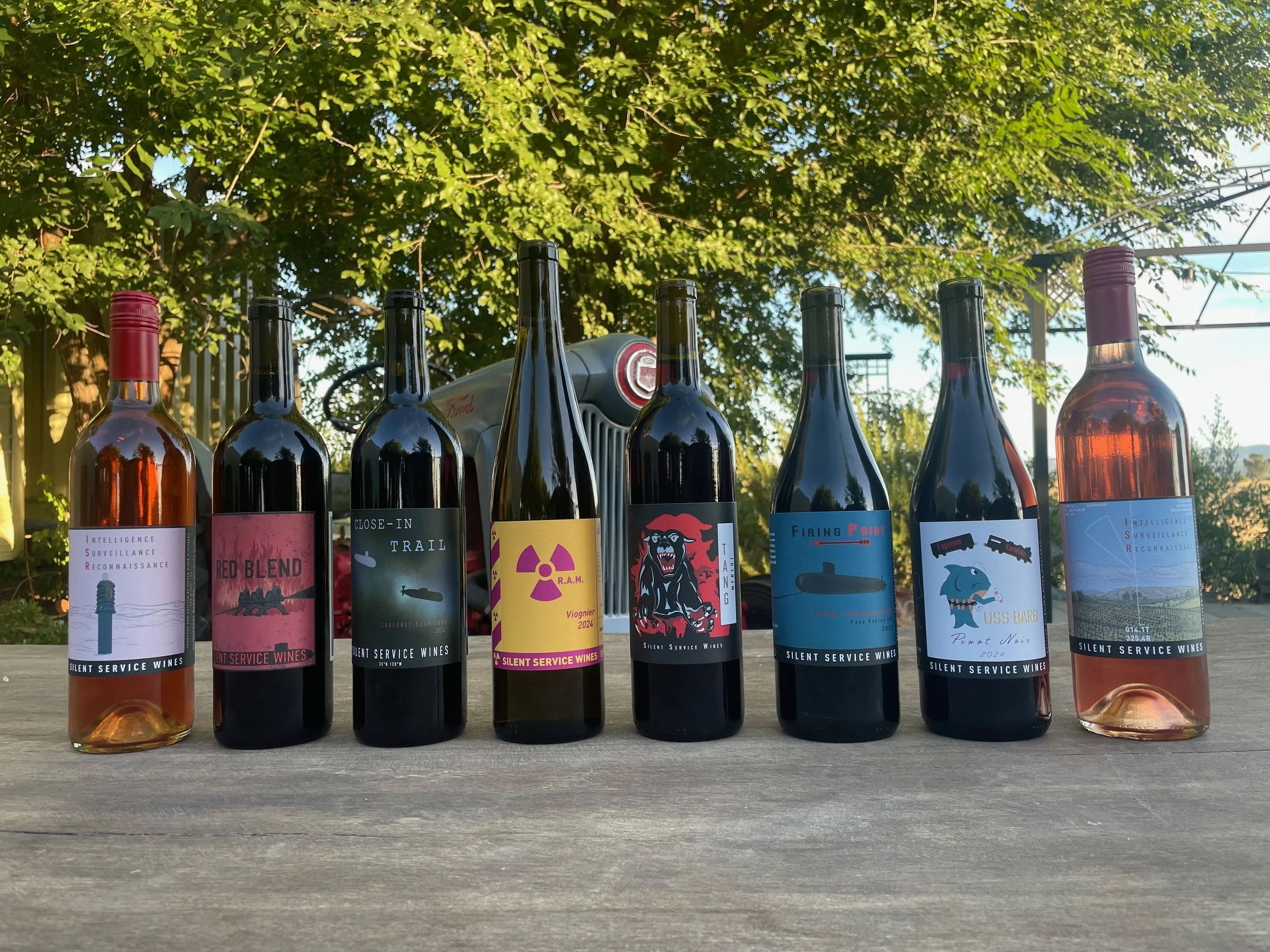 A row of nine wine bottles with colorful labels, set on a wooden surface outdoors with a background of green leafy trees and a blue sky.
