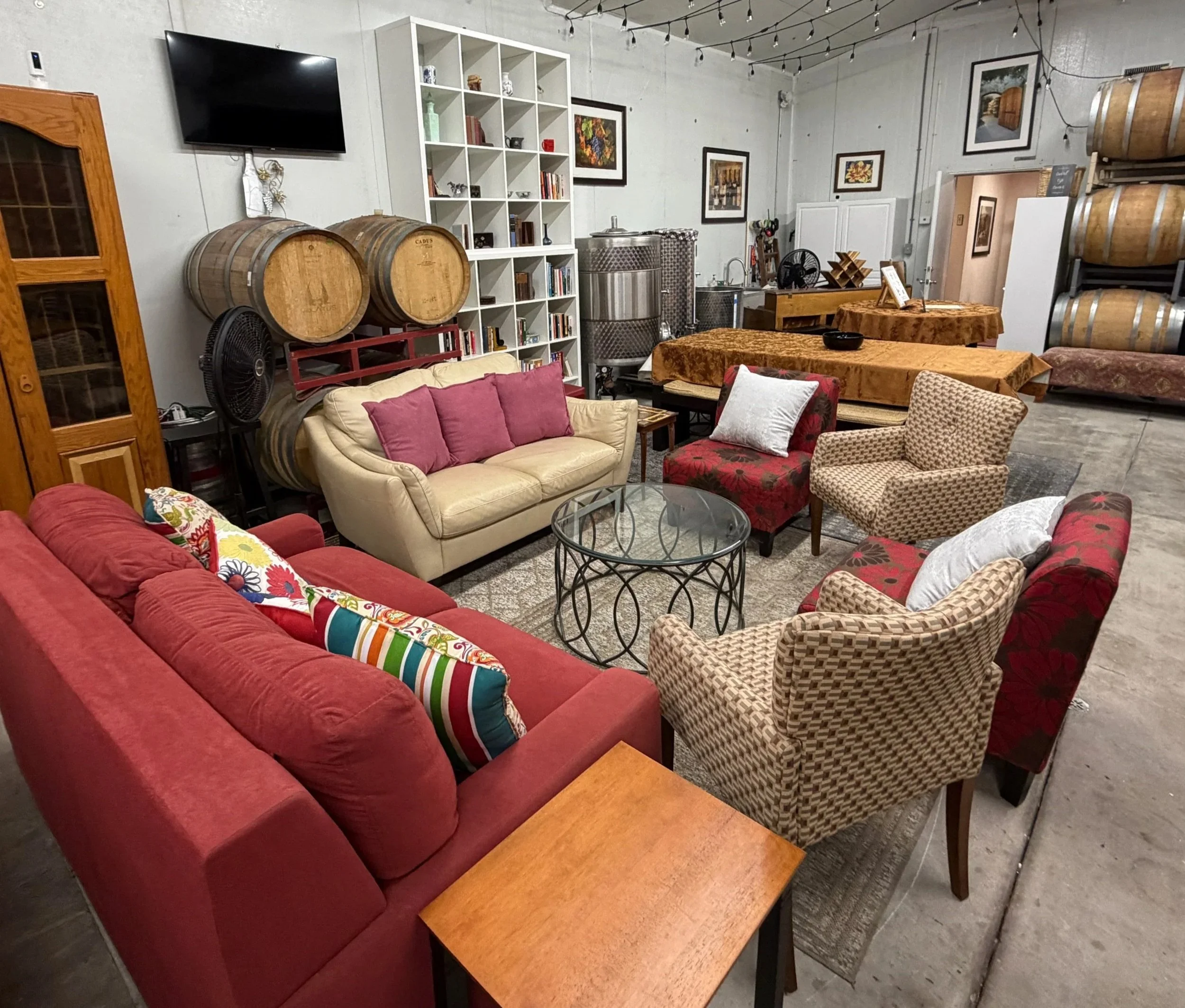 The image shows a cozy lounge area with red and patterned couches, a coffee table, and a variety of chairs. There is a white bookshelf filled with books and decorative items, a TV mounted on the wall, and several wine barrels stacked on metal racks. String lights hang from the ceiling, creating a warm ambiance.