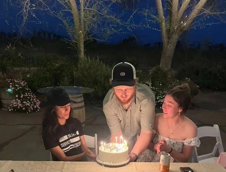 A man is blowing out candles on a birthday cake at a celebration with two women, one on each side. One woman is wearing a black hat and a black shirt, and the other woman has her hair in a bun and is smiling. The scene is outdoors at night with trees and flowers in the background.