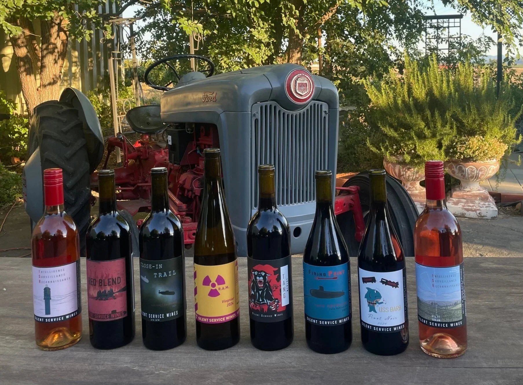 A row of eight wine bottles with various labels, placed on a wooden surface outdoors. In the background, there is a vintage Ford tractor, green plants, trees, and garden decorations.