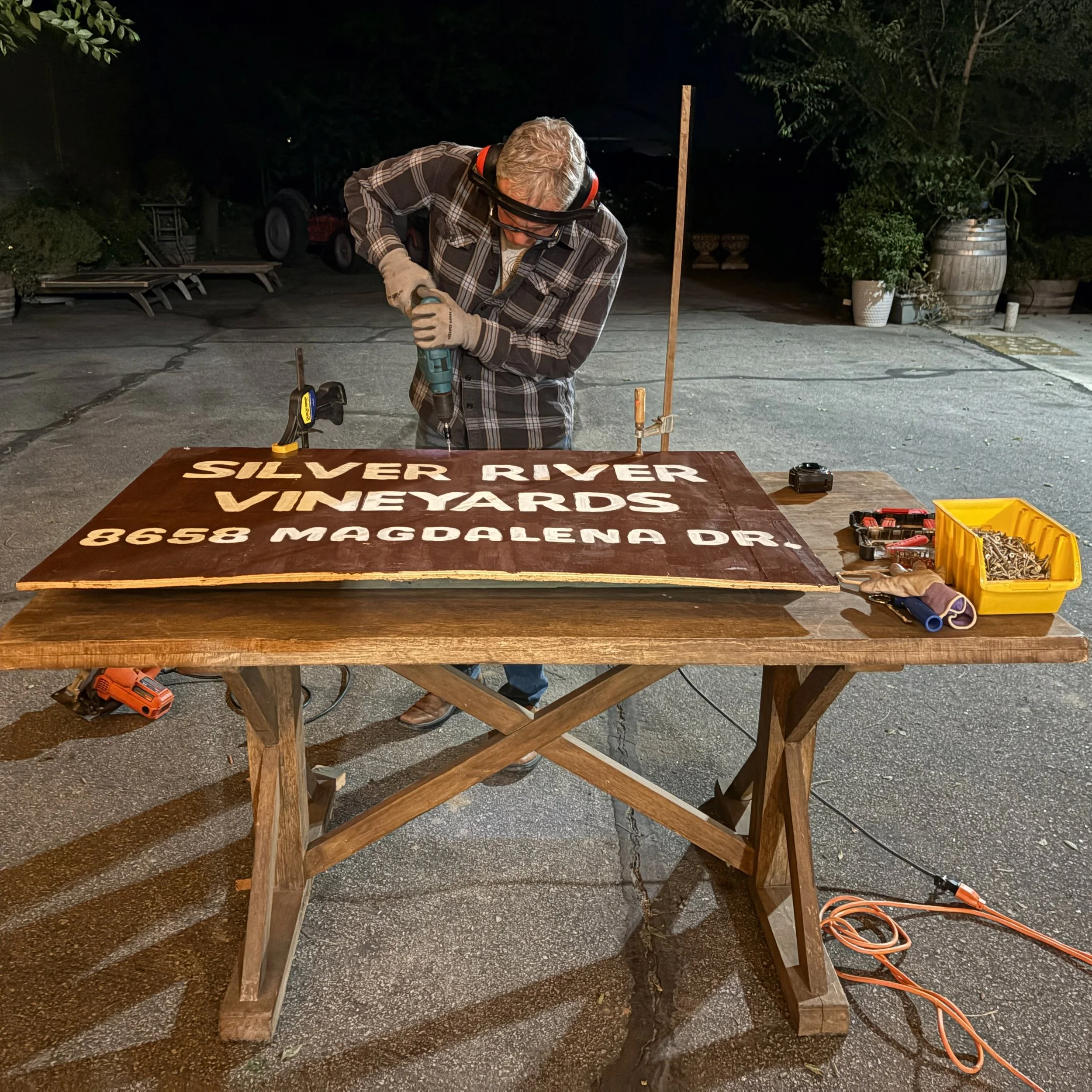 A man working outdoors at night, wearing headphones and safety glasses, using a power drill on a wooden sign that reads 'Silver River Vineyard 8658 Magdalen Dr.' surrounded by tools and equipment.