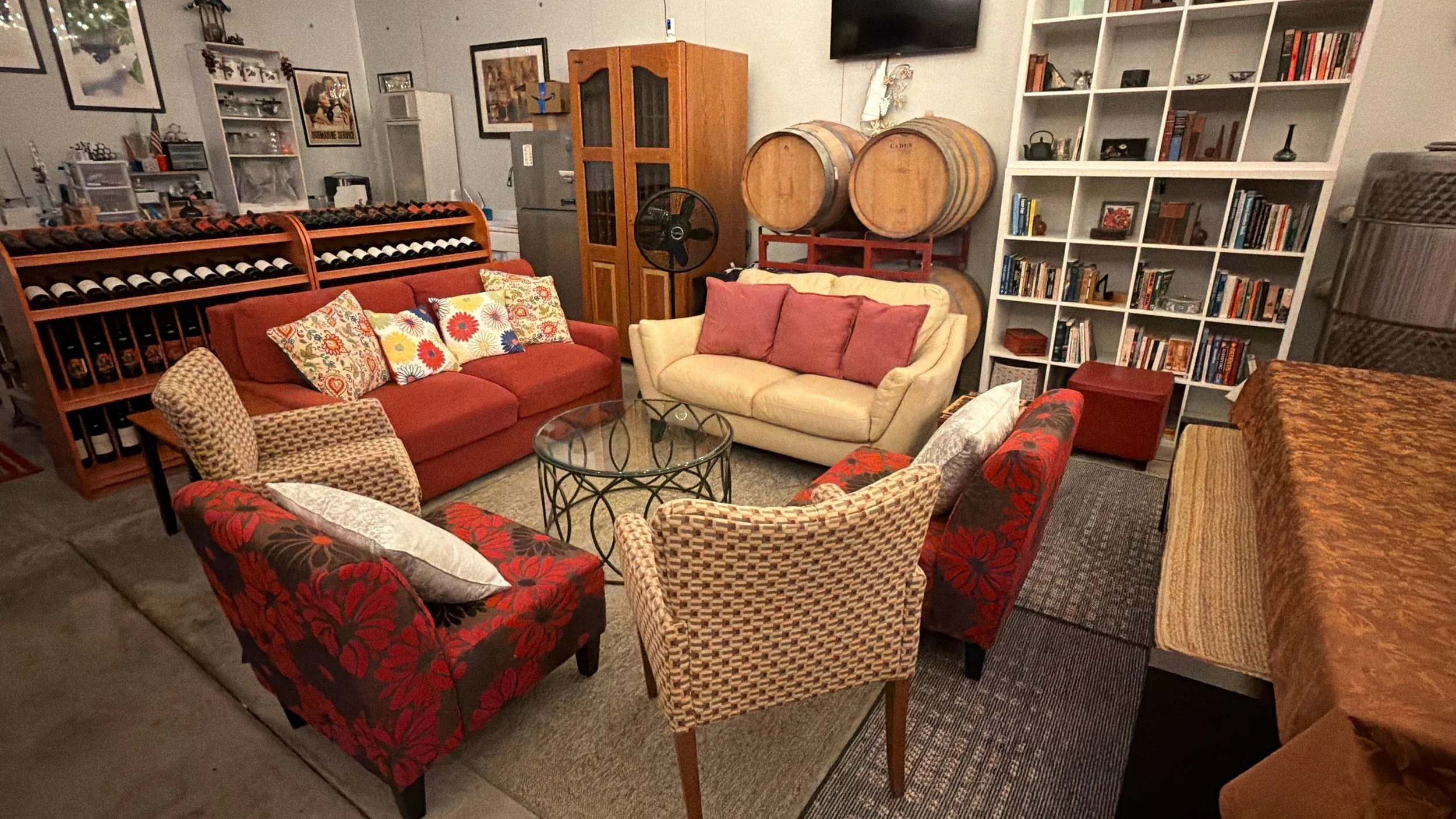 Indoor lounge with various patterned and solid color couches, a glass coffee table, a white bookshelf filled with books and decorative items, wine barrels, and framed pictures on the wall.