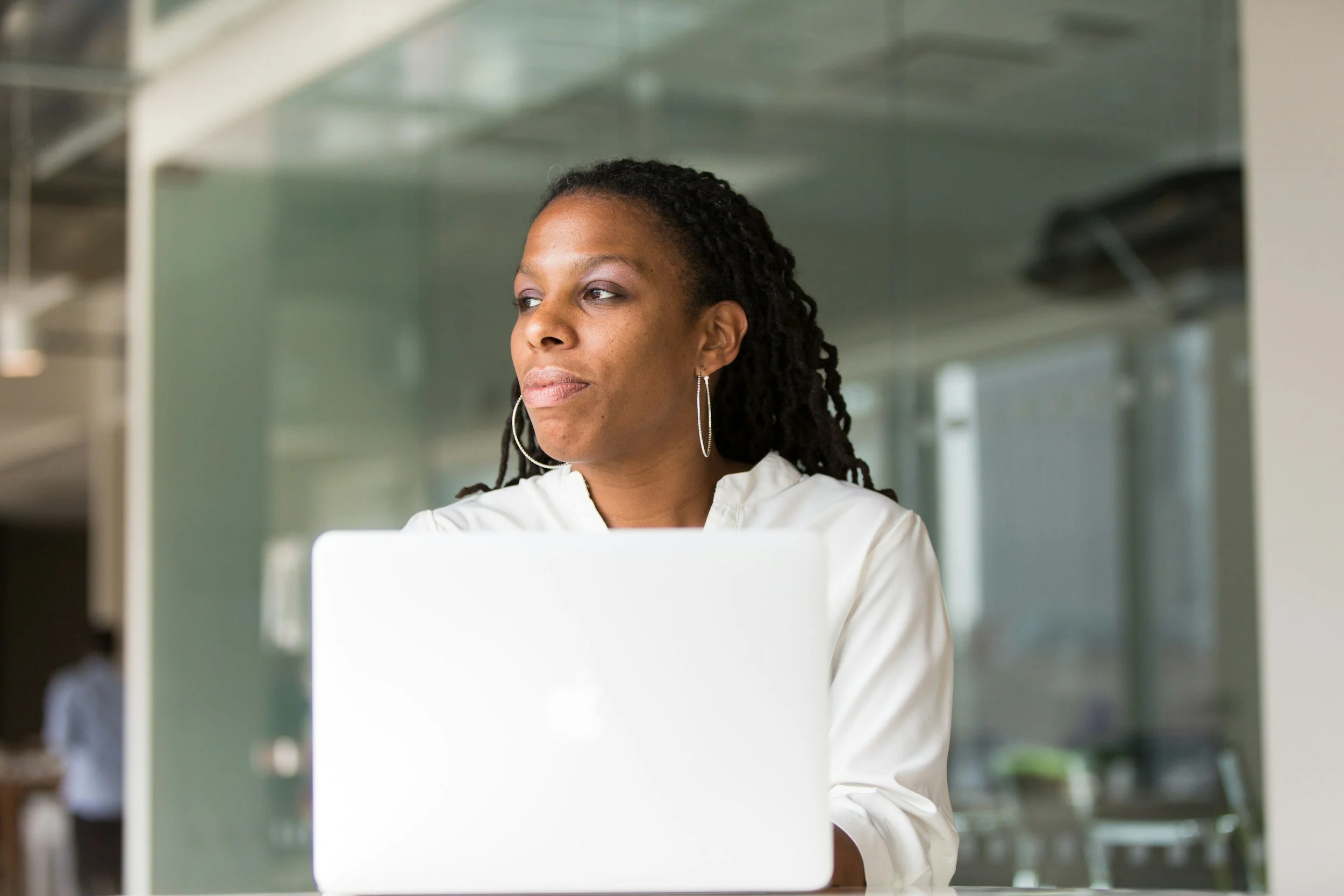 A woman with dark skin and dreadlocks wearing hoop earrings and a white top, working on a laptop in a modern, bright office space with glass walls.