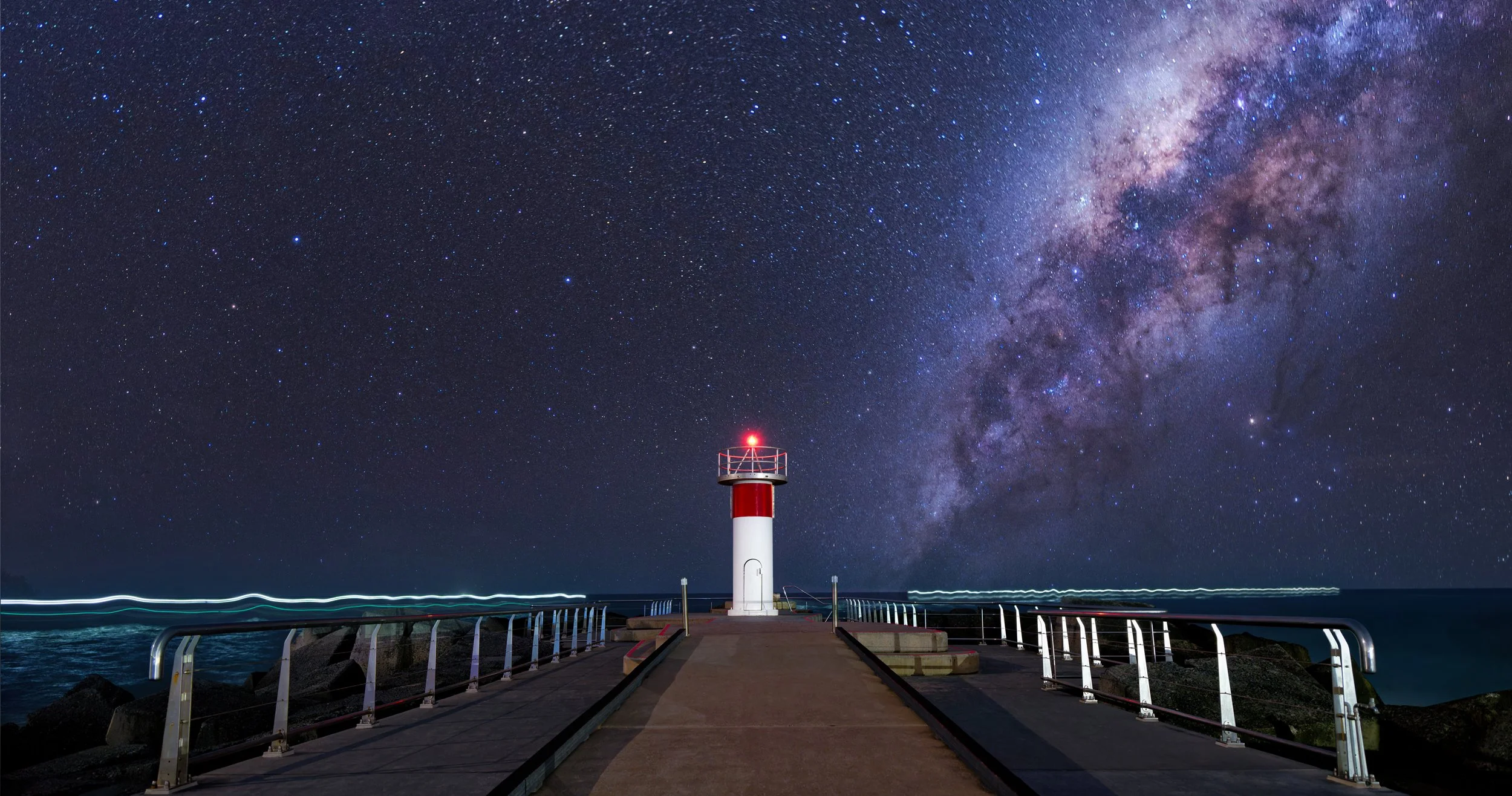Nighttime view of a lighthouse on a pier, with a star-filled sky and the Milky Way galaxy overhead.