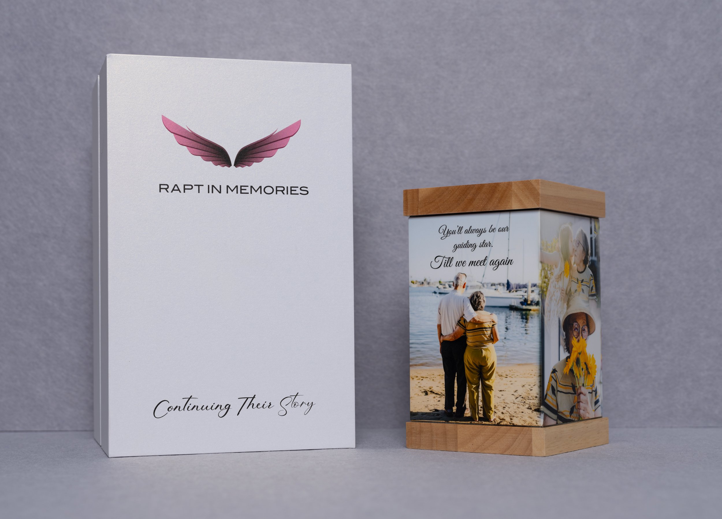 A white box with a pink butterfly logo and the text "RAPT IN MEMORIES" and "Continuing Their Story" next to a decorative wooden box featuring a photo of an elderly couple walking by a harbor with boats, accompanied by the quote "You’ll always be our guiding star. Till we meet again."