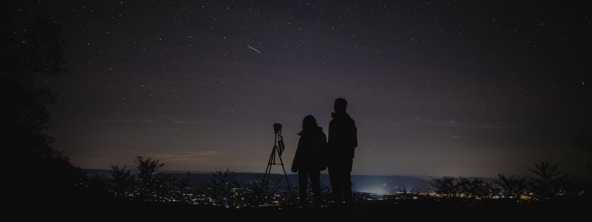 Silhouettes of two people and a camera on a tripod watching stars in the night sky over a cityscape.