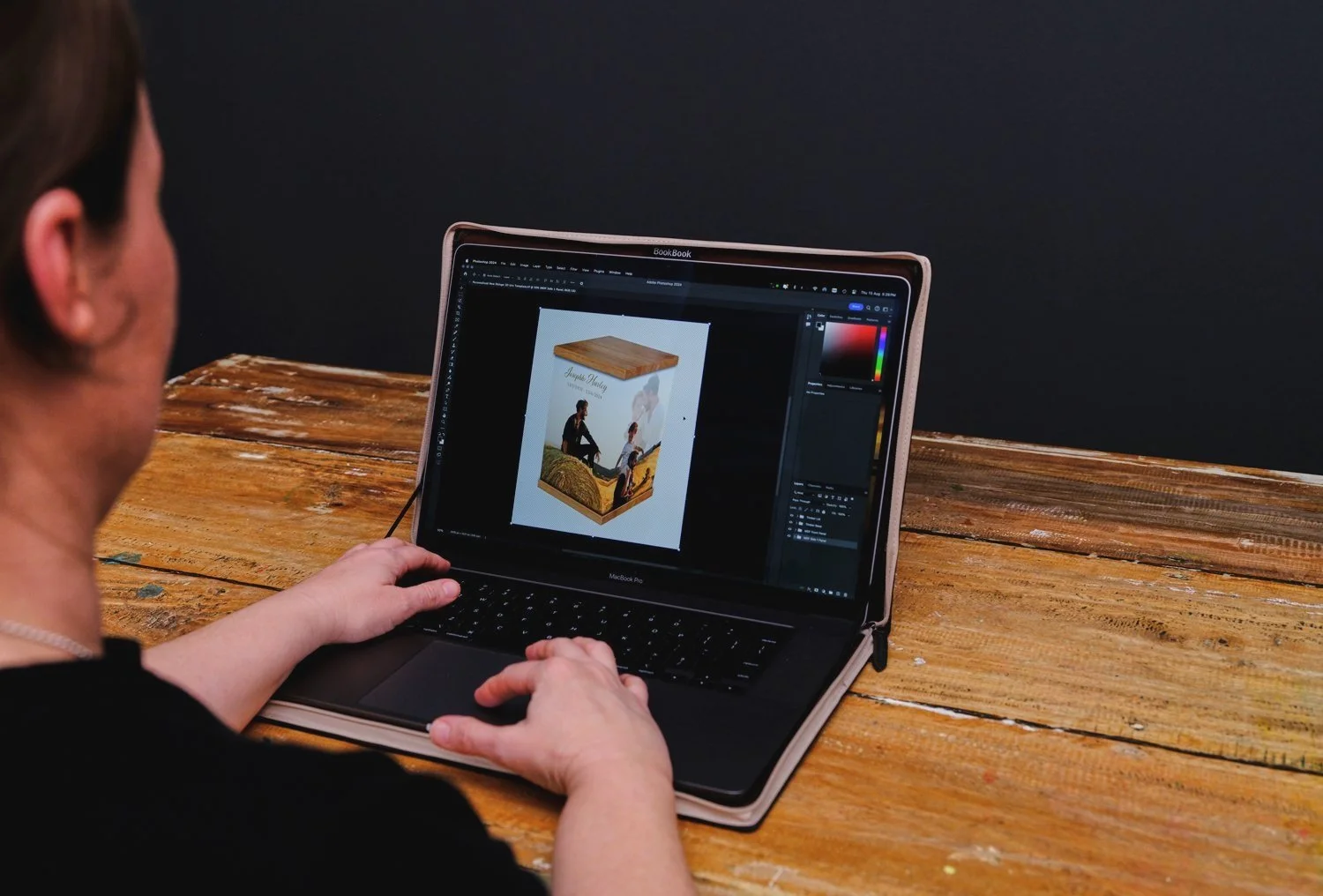 Person working on a MacBook Pro, designing a book cover with an image of two people sitting on a hay bale and a guitar, displayed on the screen. The laptop is on a rustic wooden table.