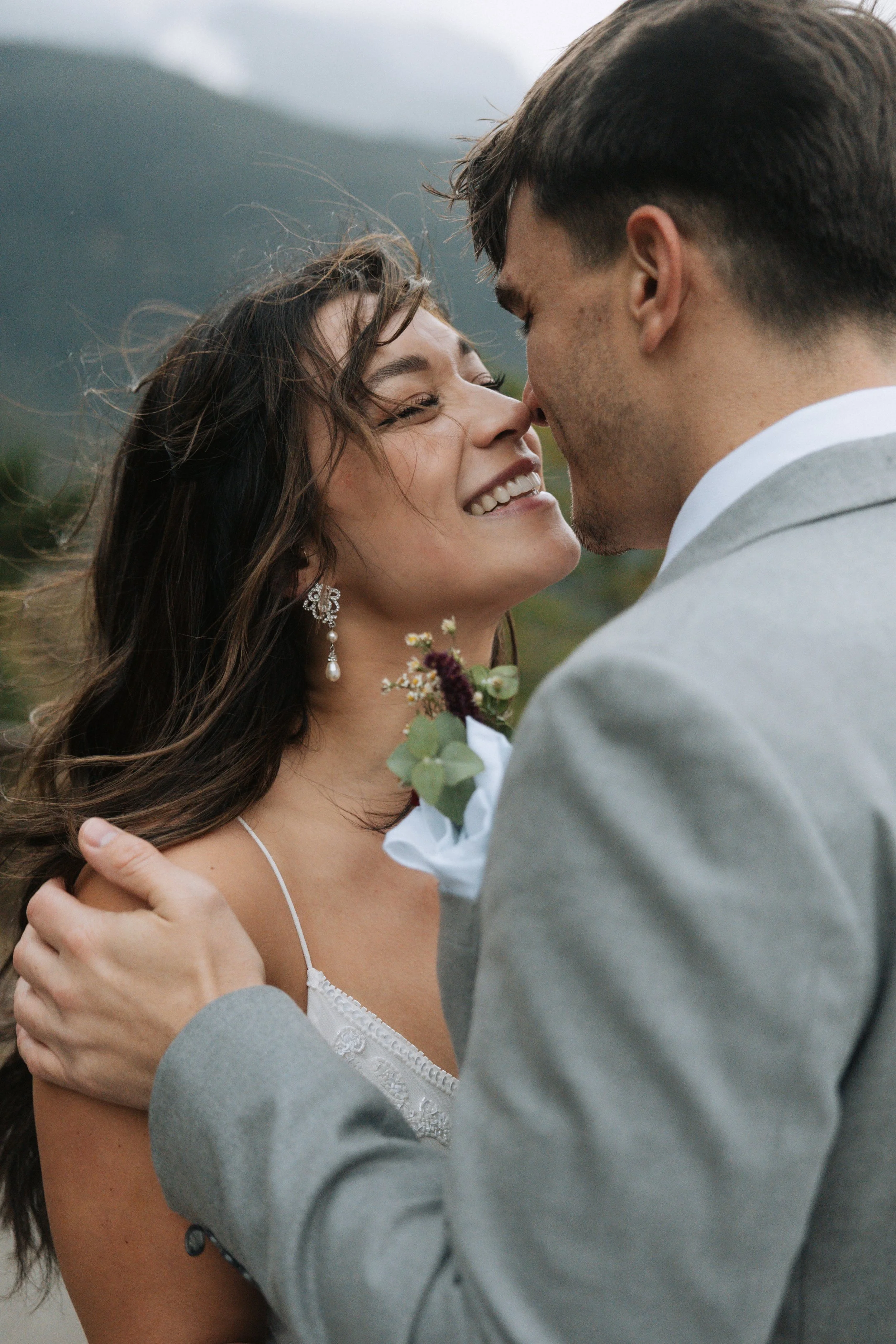 Bride smiling as her groom kisses her outdoors with mountains in the background