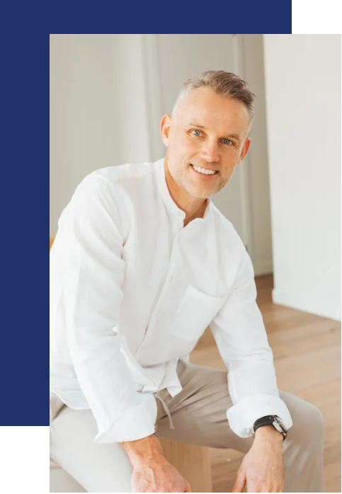 A smiling middle-aged man with gray hair and a beard, wearing a white shirt and khaki pants, sitting on the floor in a bright room with hardwood floors and a white wall.