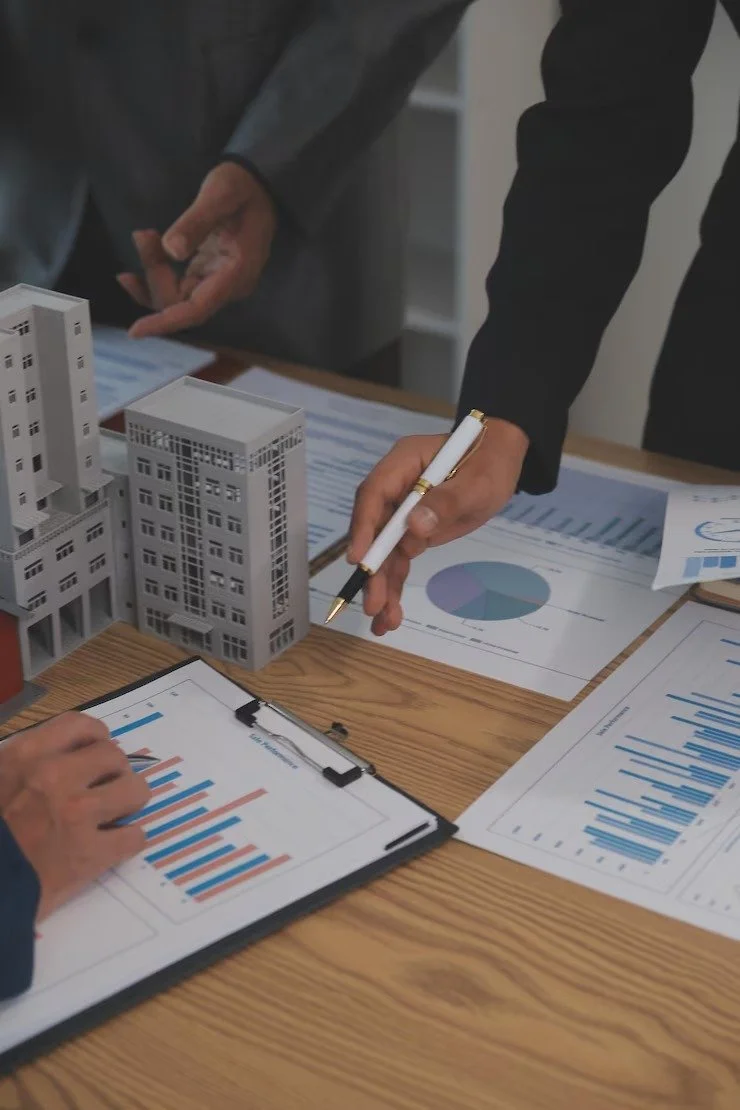 Business professionals discussing architectural models and analyzing data charts on a wooden table.