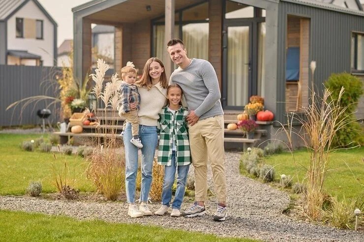 Family of four, including a man, woman, and two young girls, standing outside their modern house decorated with fall pumpkins and gourds in the yard.