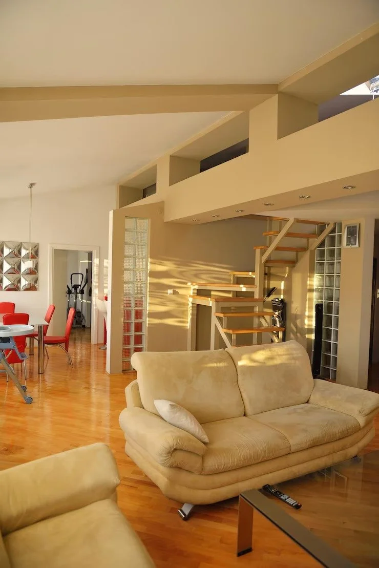 Living room with beige sofas, wooden staircase, glass block wall, red dining chairs, and exercise equipment in the background.