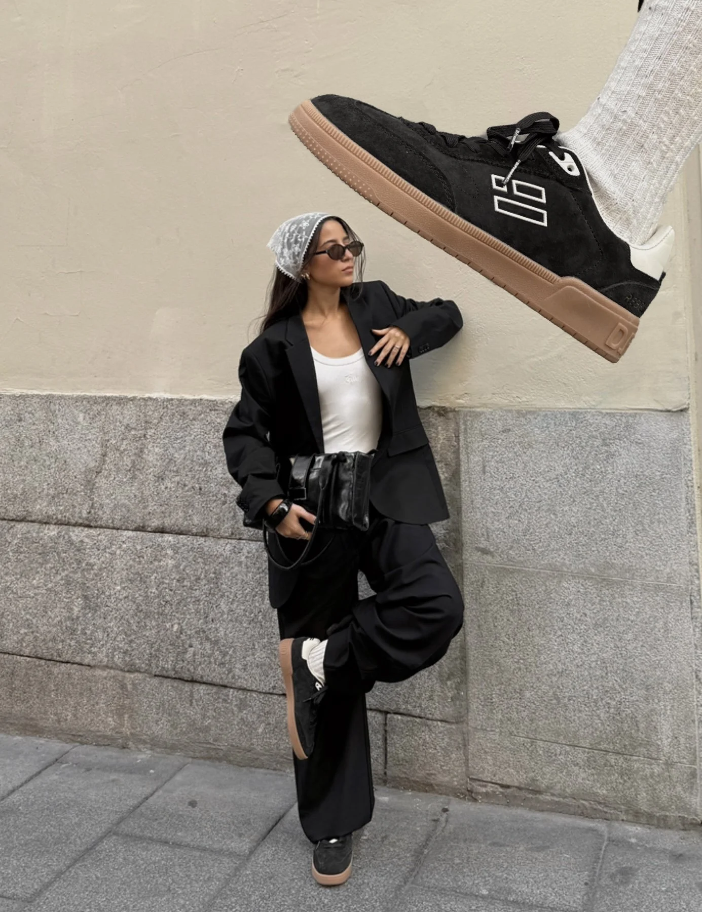 Mujer con ropa negra, gafas y sombrero de encaje, posando contra una pared de piedra, con un pie elevado con un zapato deportivo hacia la cámara.
