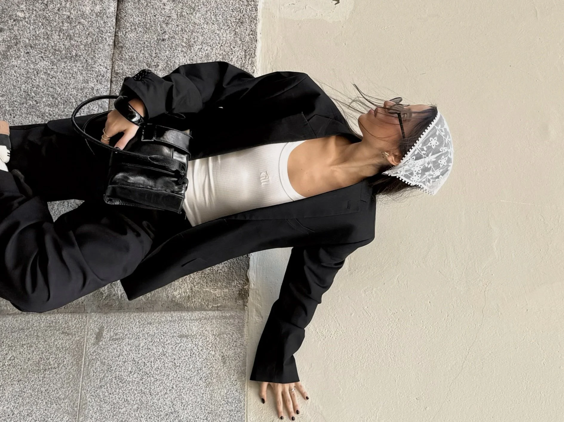 Mujer con gabardina negra, camiseta blanca y falda corta, usa gafas y sobre la cabeza lleva una cofia blanca con bordado de flores, lleva bolso negro y se apoya en el suelo con una mano.