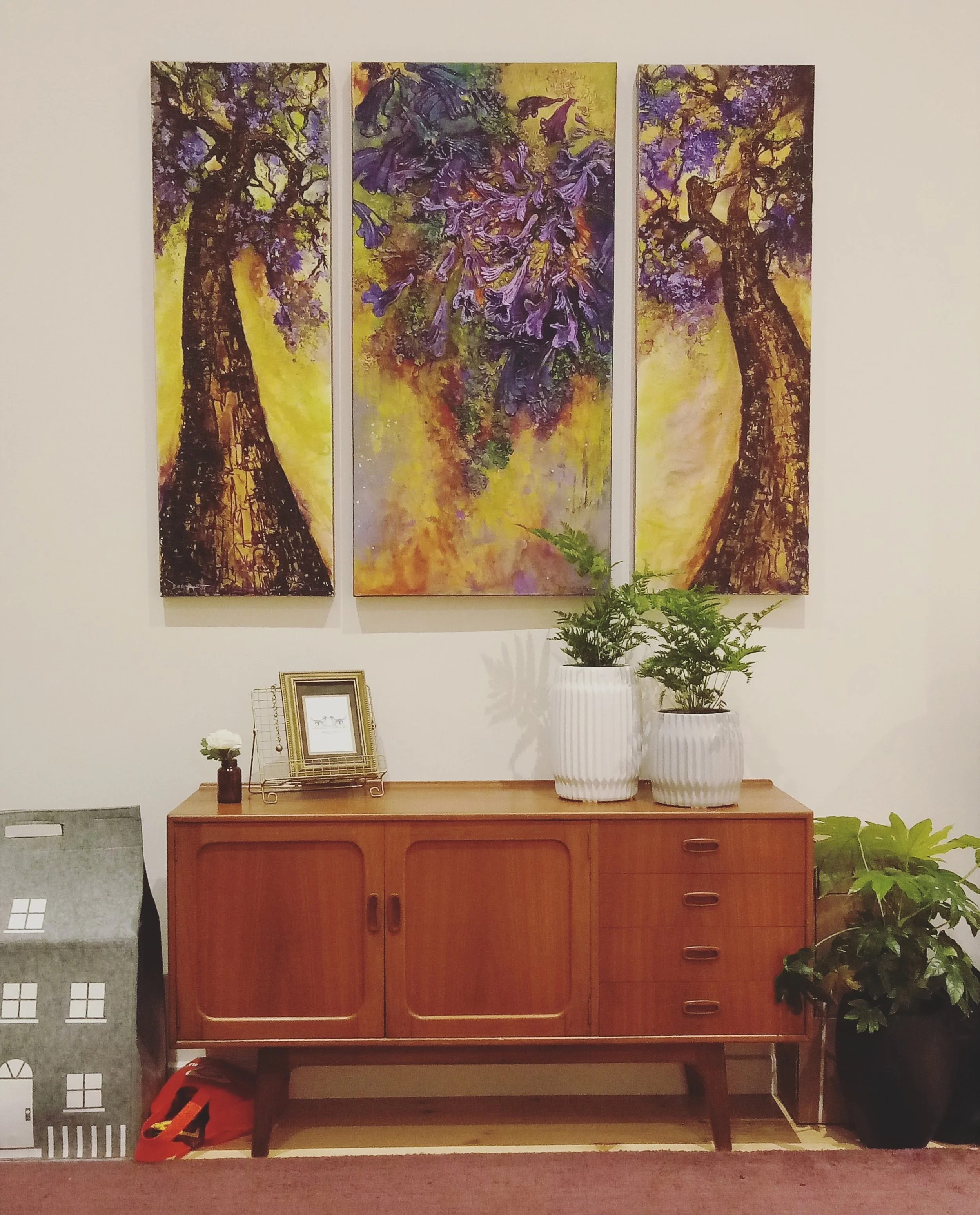 A wooden sideboard with decorative items, and two potted plants on top, against a wall with three vertically oriented abstract purple and yellow paintings of trees.