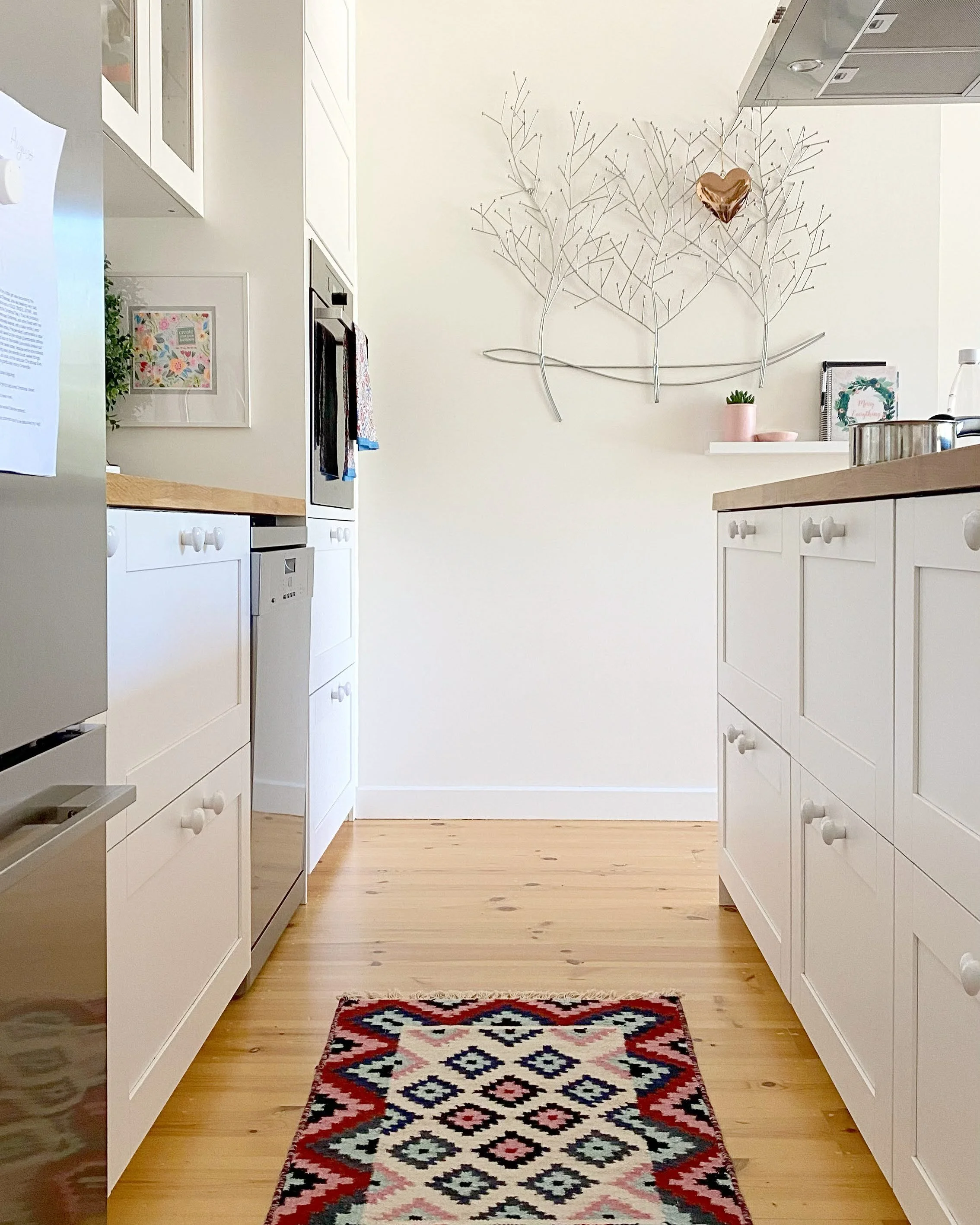 Kitchen with white cabinets, wooden floor, a colorful geometric rug, and wall decorations including a heart-shaped foil balloon and a framed floral pattern.