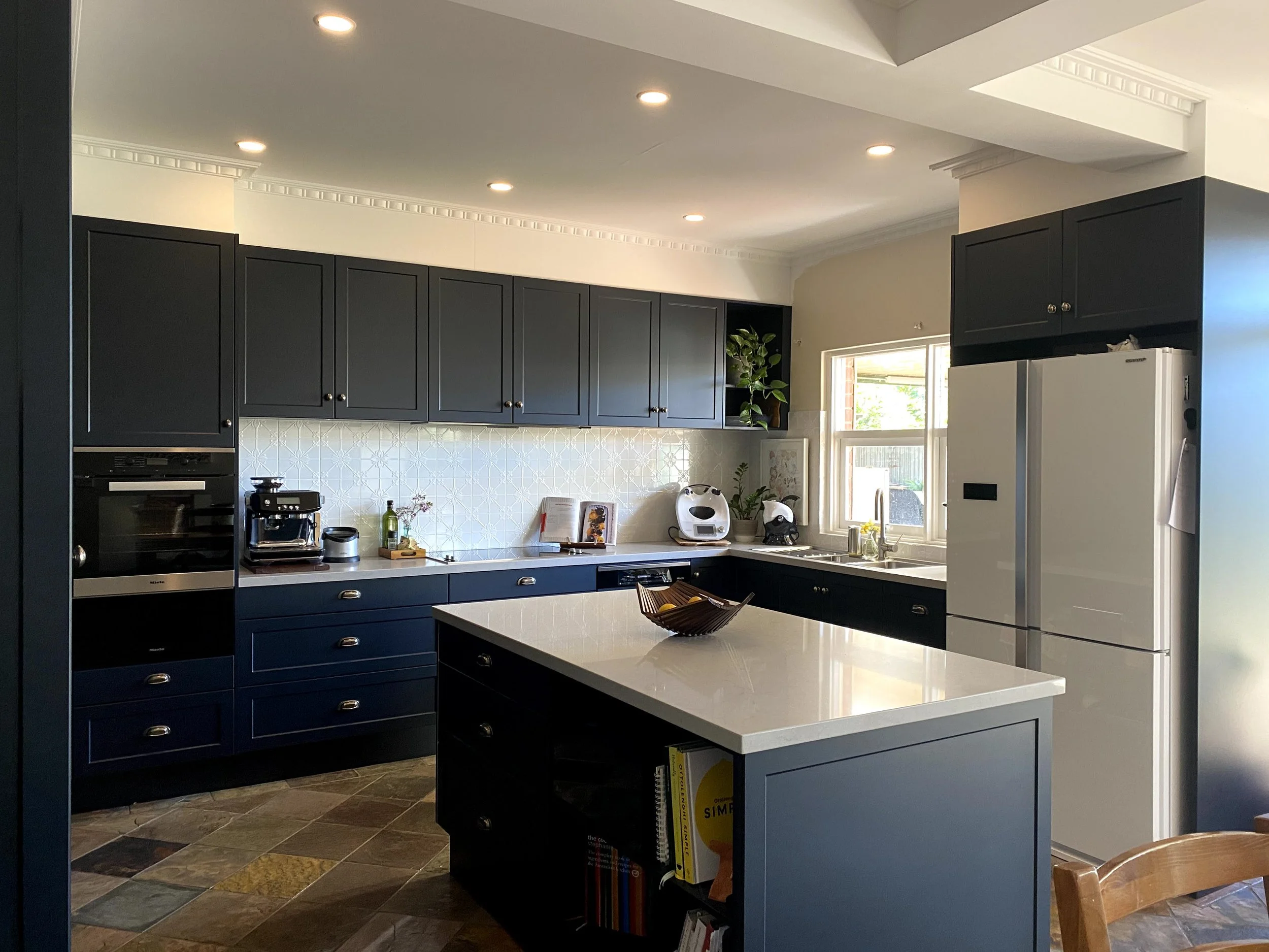 A modern kitchen with dark blue cabinets, white countertops, and a white tile backsplash. There is a window letting in natural light. Appliances include an oven, a coffee machine, and a white refrigerator. The floor is tiled with multicolored slate tiles.