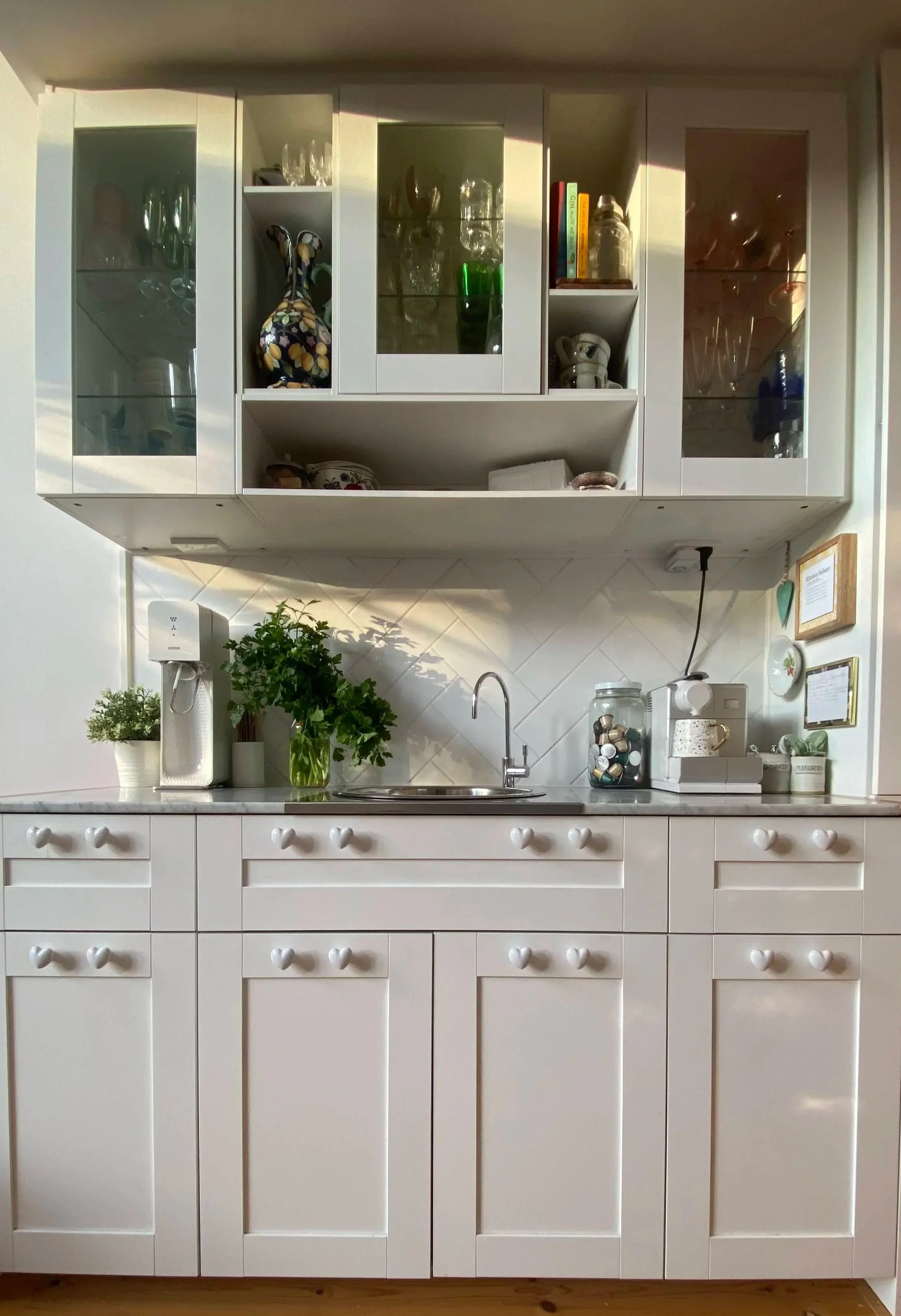 White kitchen cabinet with glass doors and heart-shaped knobs, plants on the countertop, and kitchen appliances including a coffee machine and jars, wall-mounted with framed pictures.