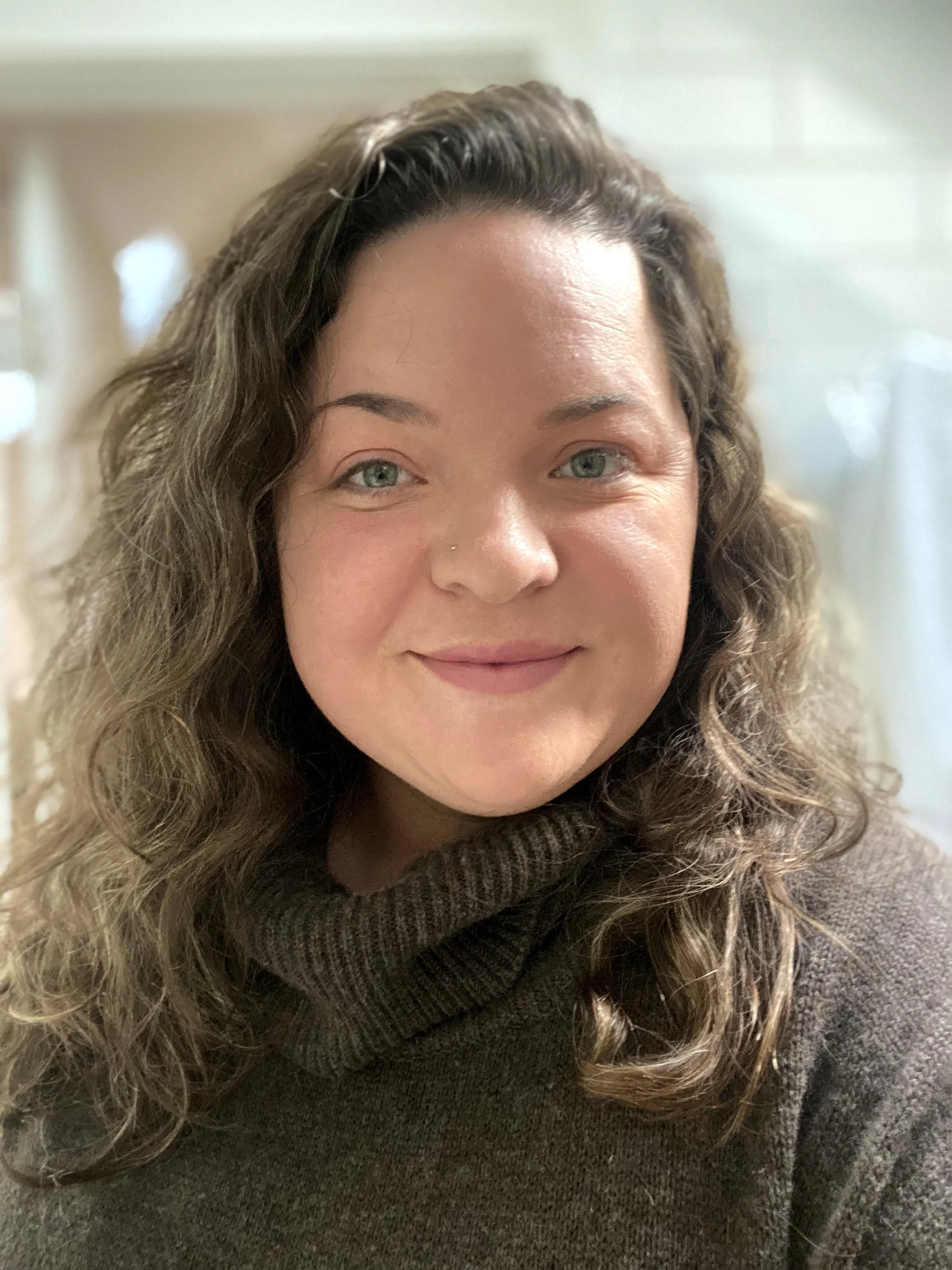 Close-up of a smiling woman with curly brown hair, blue eyes, and a nose piercing, wearing a dark sweater.