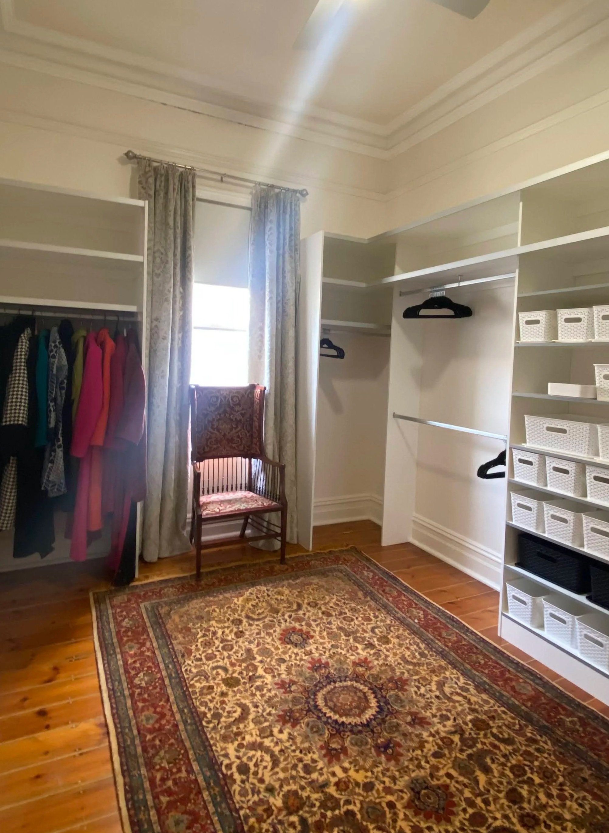A walk-in closet with open shelving, a clothing rack with various garments, a chair, a window with floral curtains, and a patterned area rug on hardwood floors.