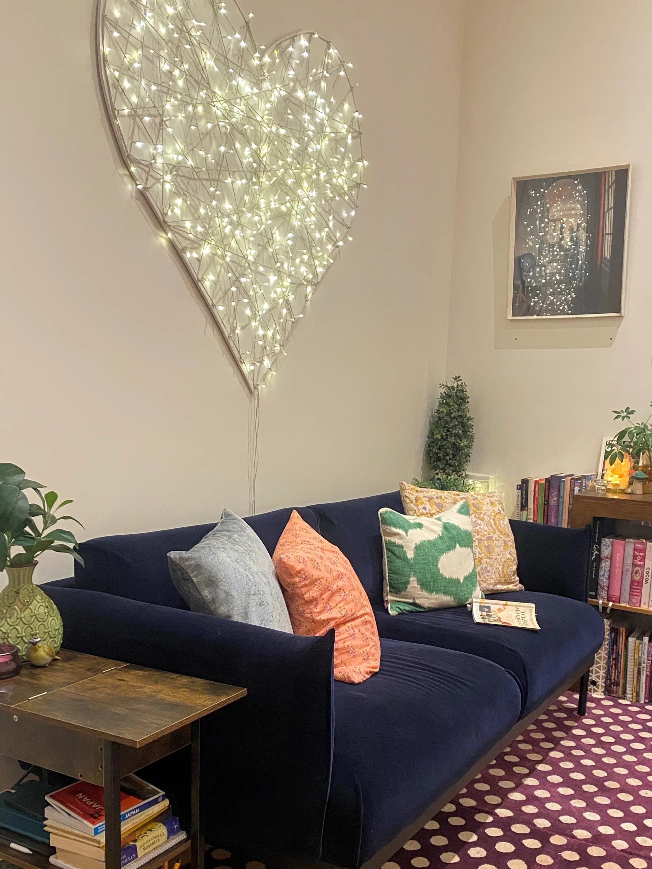 A living room with a dark blue sofa decorated with colorful pillows, a wooden side table, a bookshelf filled with books, a potted plant, and a wall-mounted mirror surrounded by string lights forming a heart shape. The room has a patterned maroon and white rug.