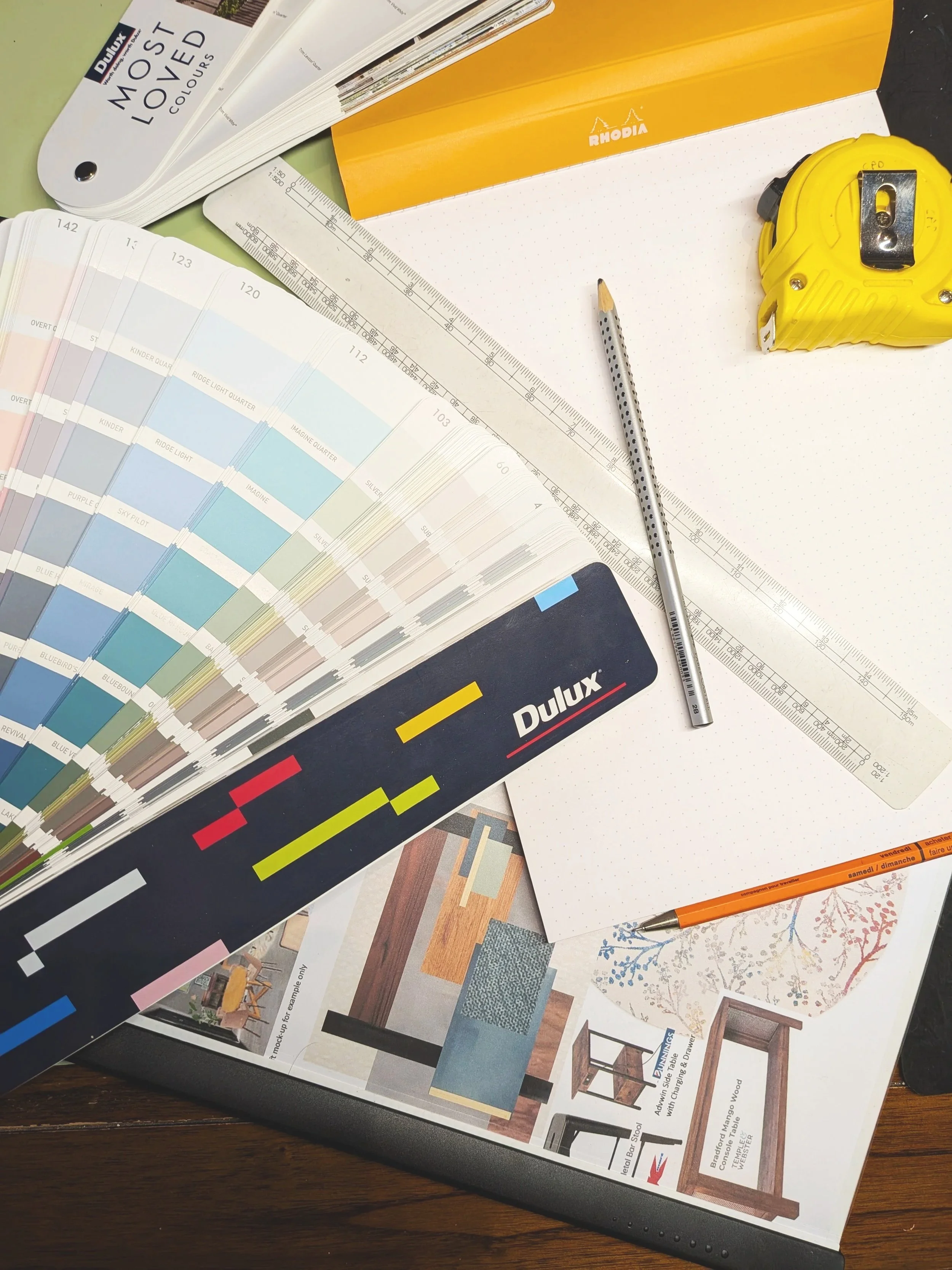 Color swatch book, a yellow measuring tape, a gray pencil with black dots, a color sample booklet, and a pen on a white dotted paper sheet.